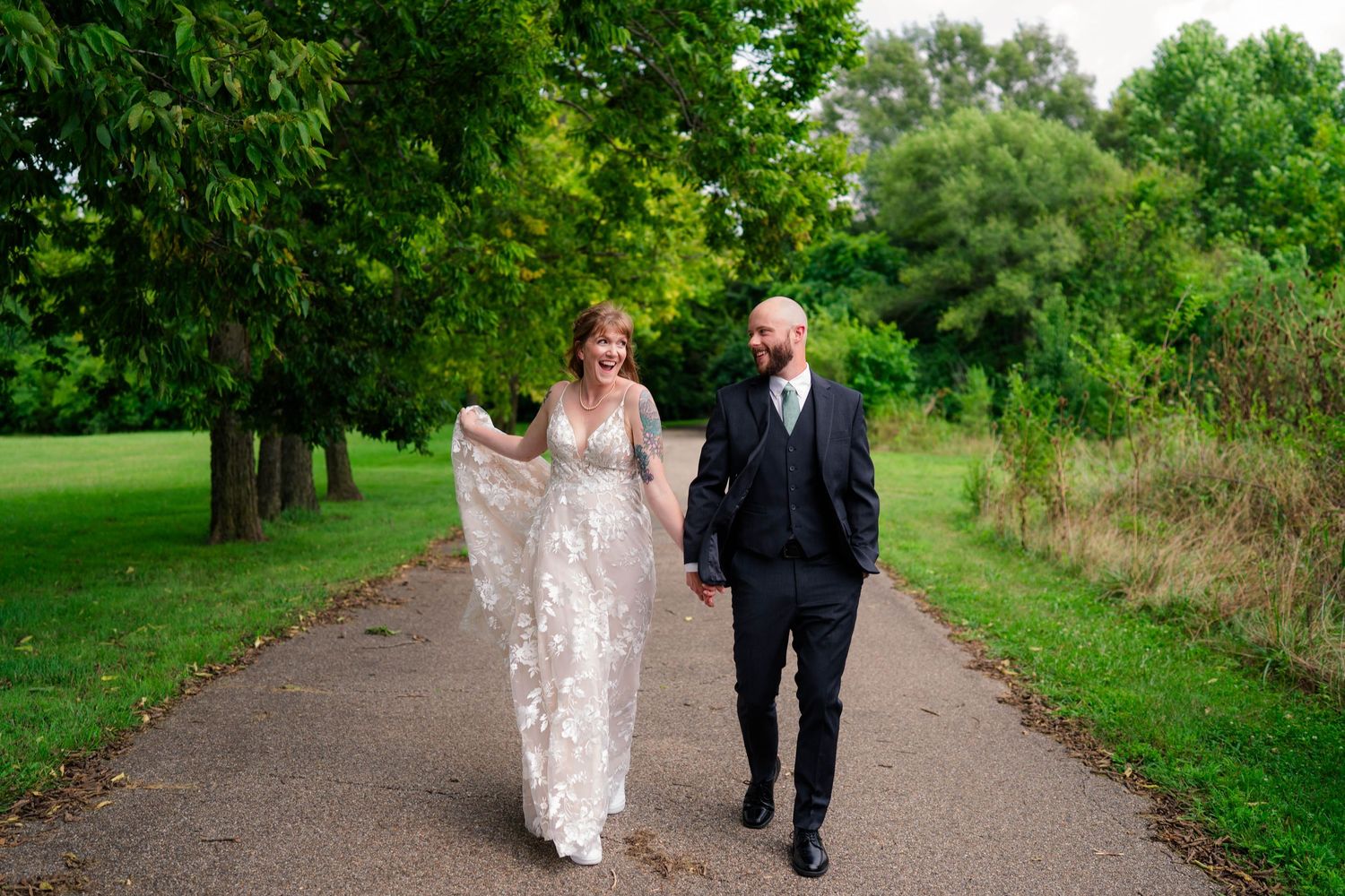 Bride and groom walking hand in hand along a lush green park pathway on their wedding day.