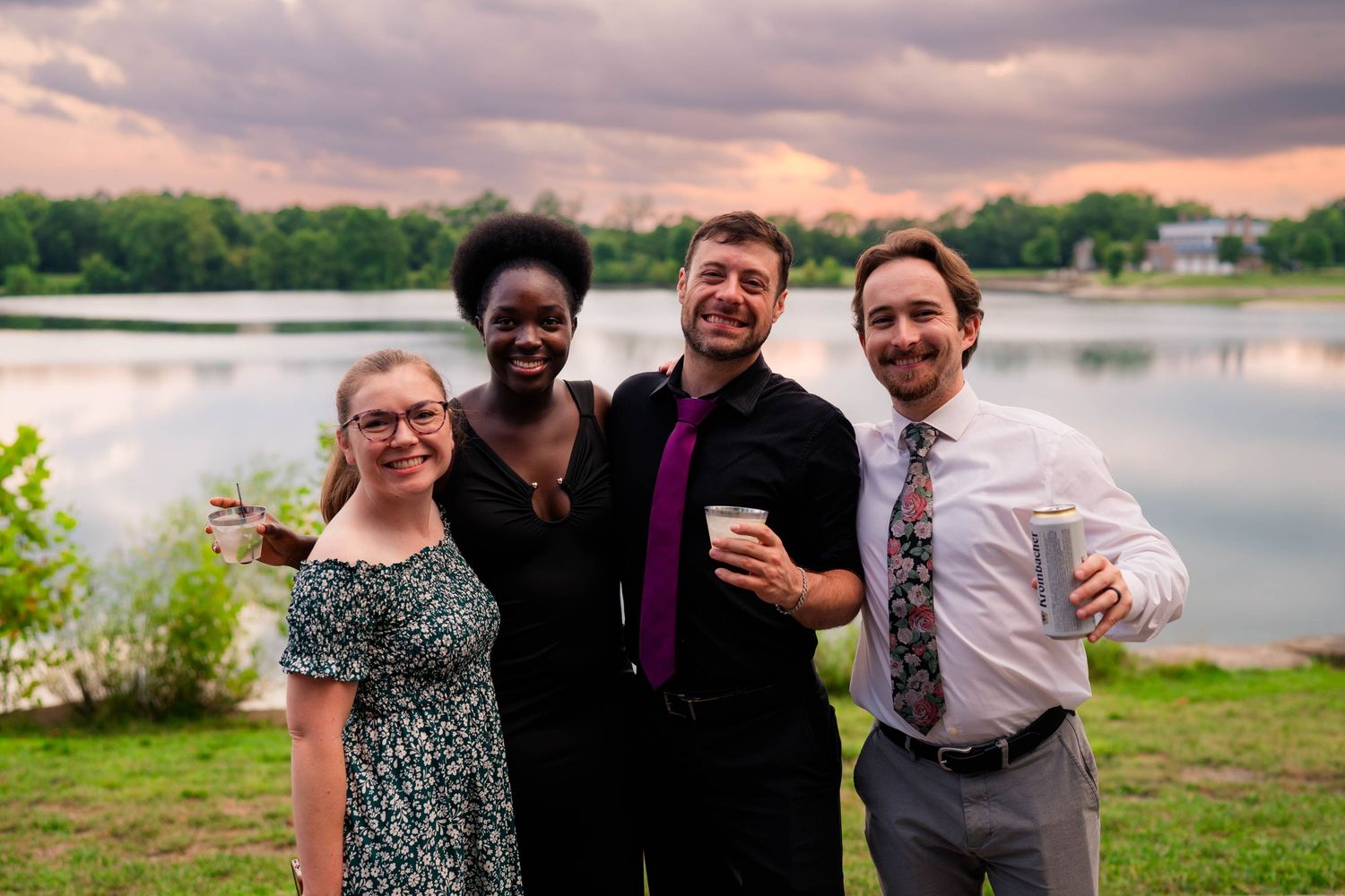 Four friends smile together outdoors near a lake at sunset during a wedding celebration.