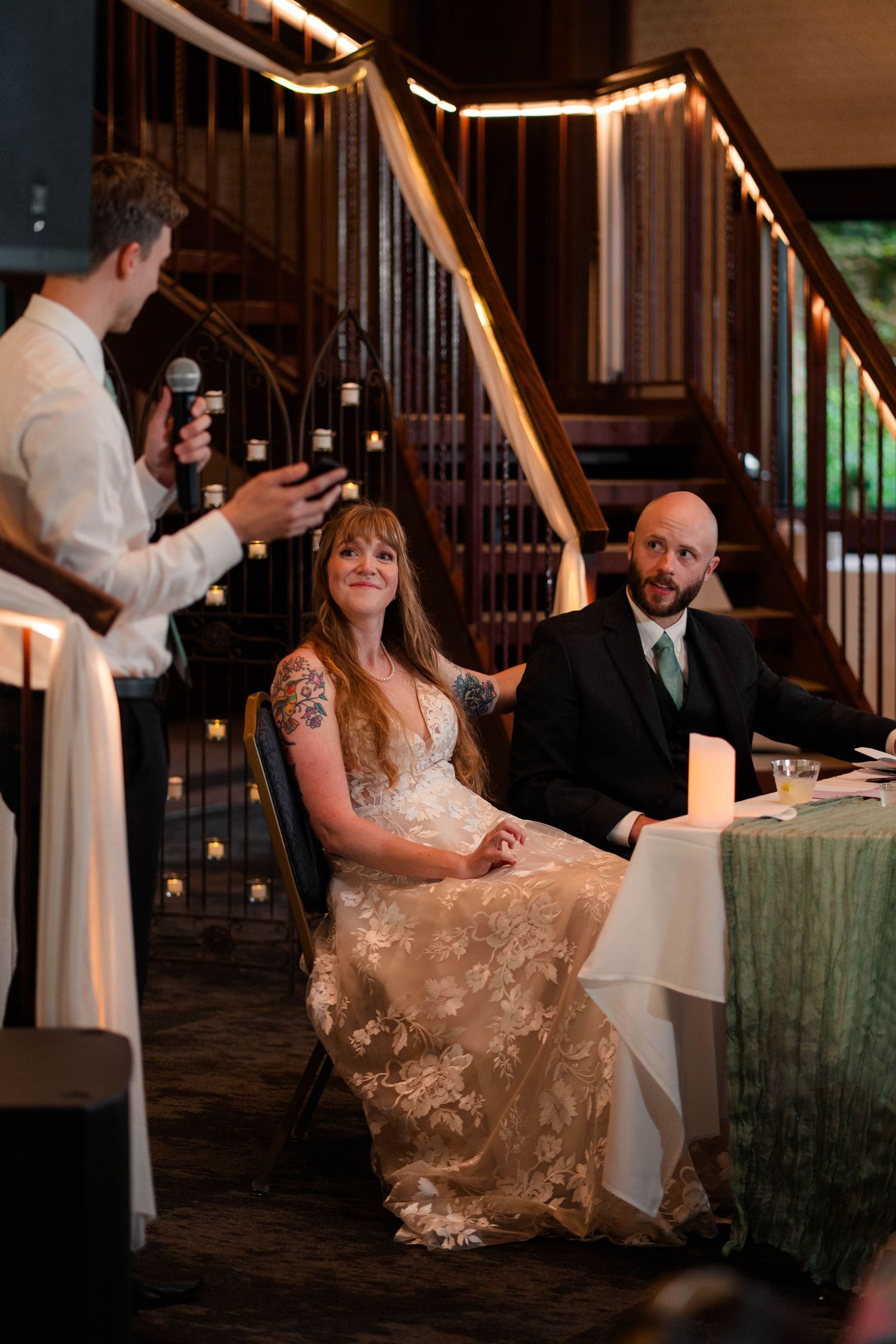 Bride and groom smile while listening to best man give wedding toast at reception.