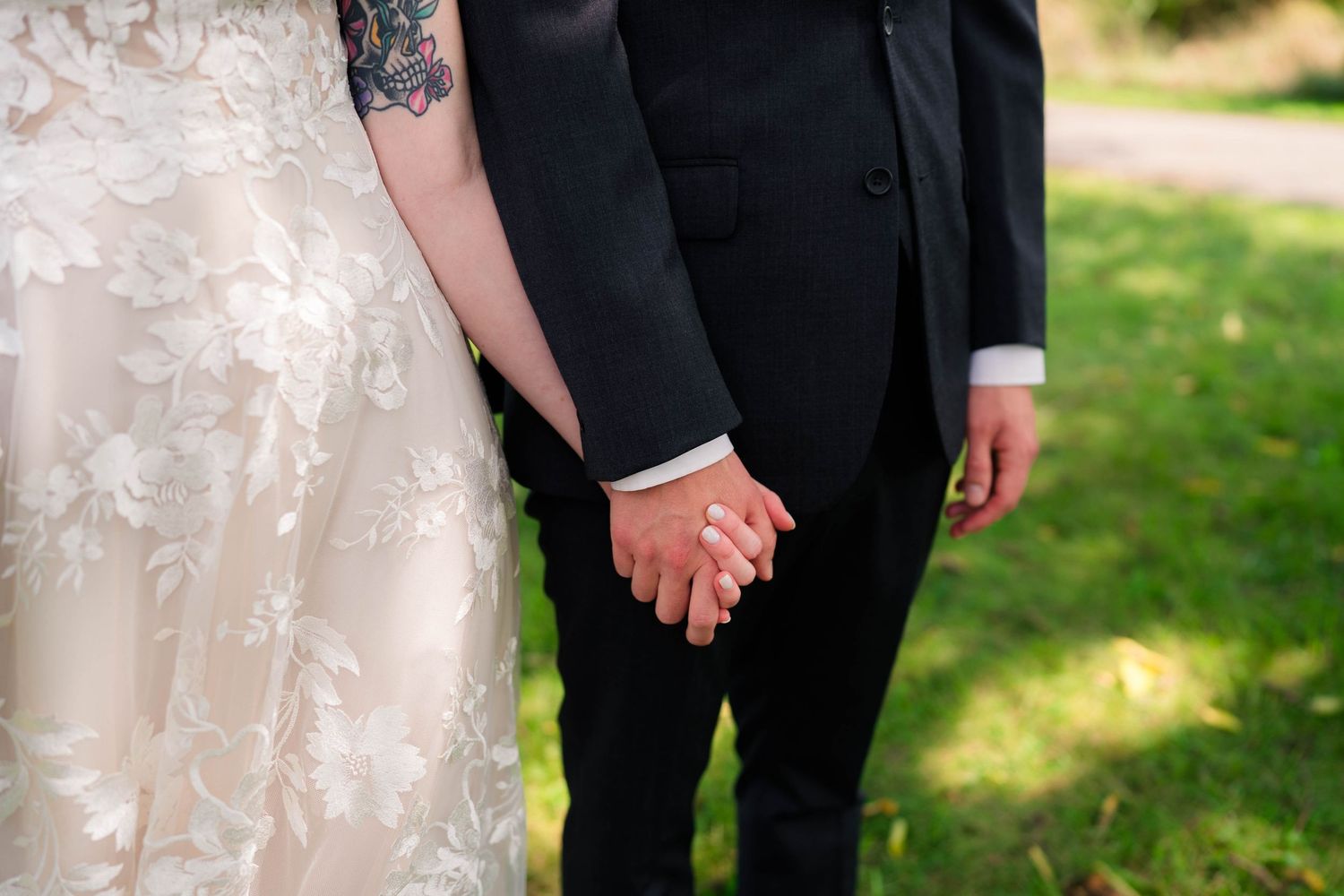 A close-up of a bride and groom holding hands, showing wedding rings and her floral lace dress.