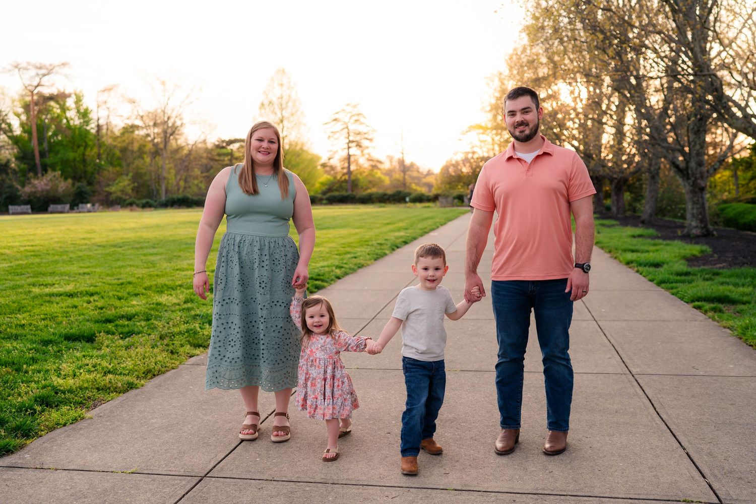 Family of four walking on a park path at golden hour, parents holding hands with two young children.