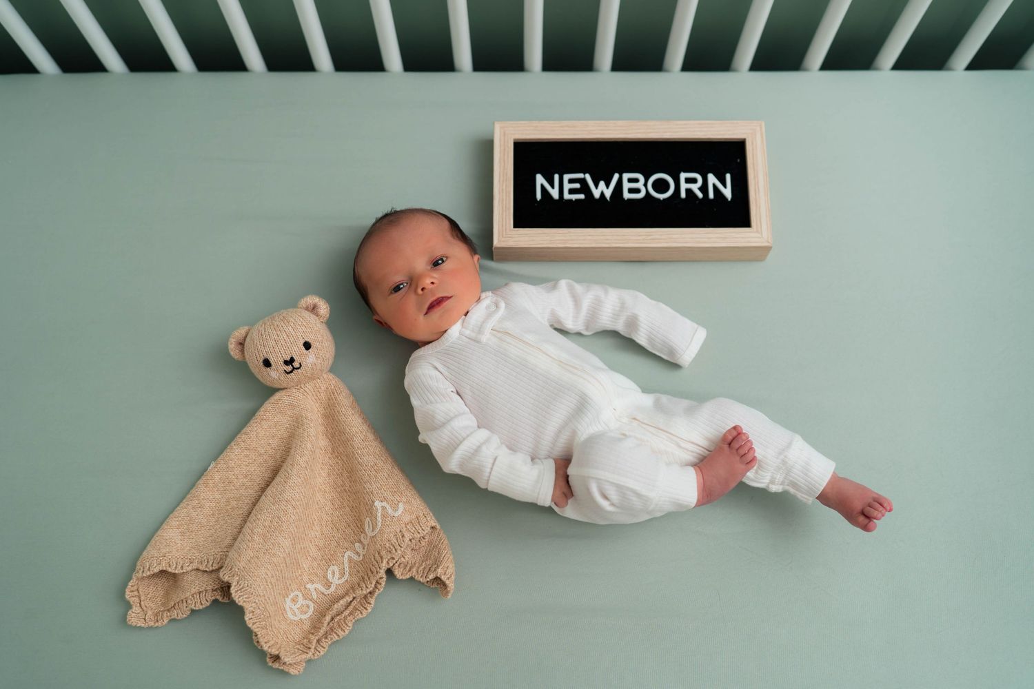 Newborn baby in white onesie lying in crib beside teddy bear lovey and chalkboard sign reading NEWBORN.