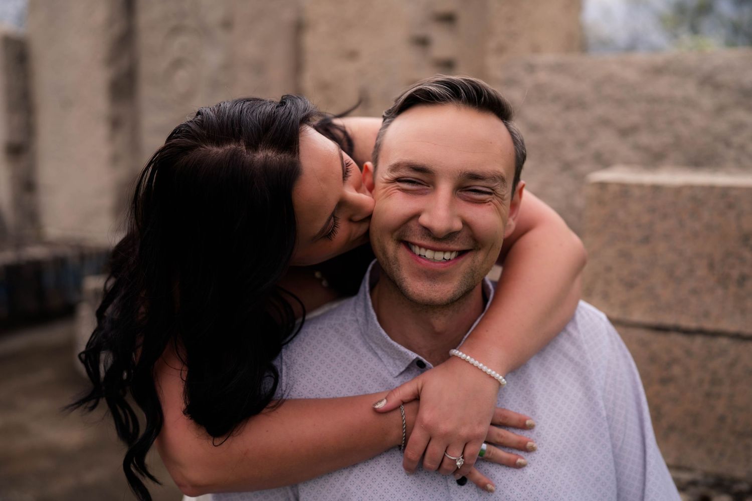 A smiling man gets a kiss on the cheek from a dark-haired woman during an outdoor engagement photo session.
