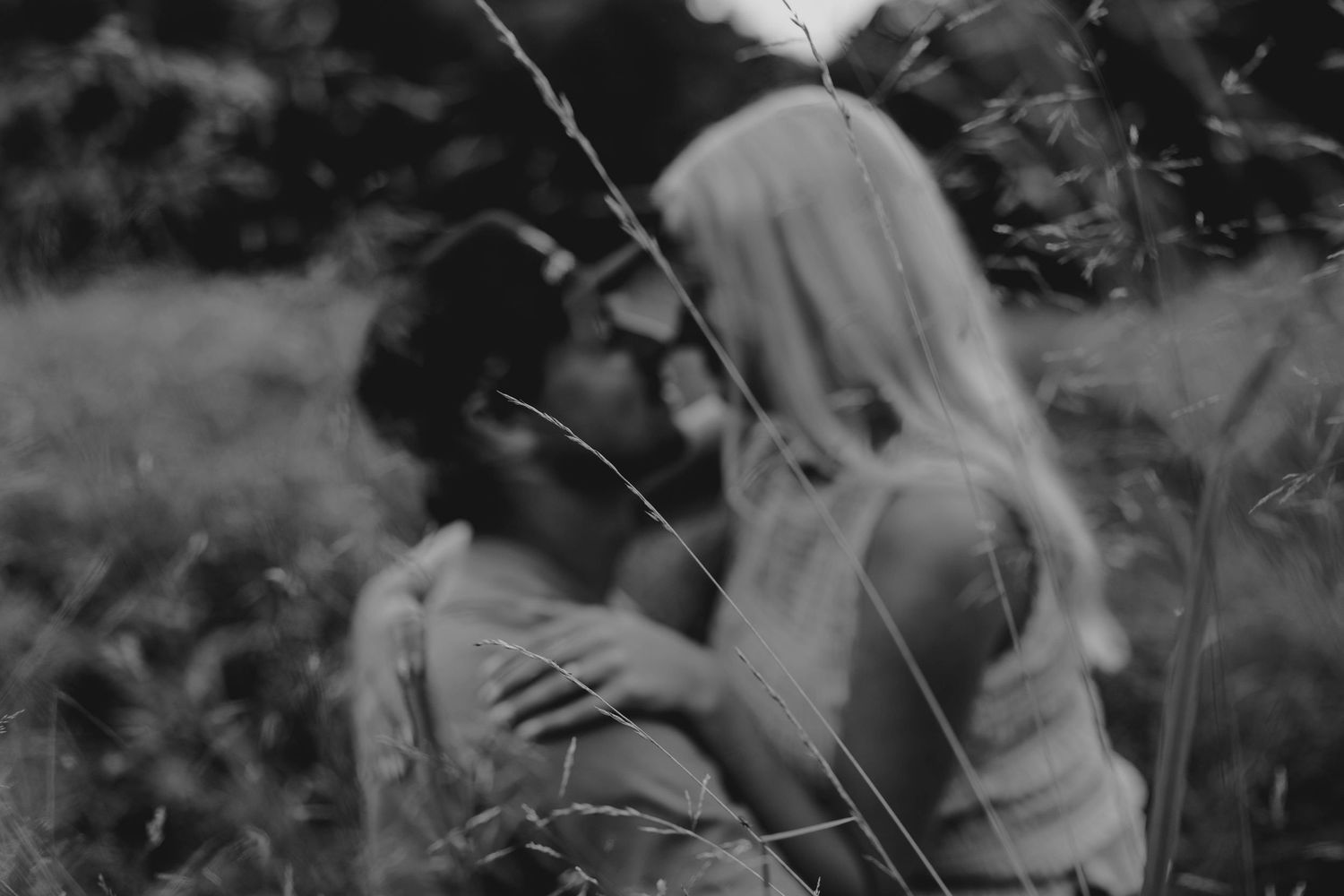 Black and white photo of a romantic couple almost kissing in a field of tall grass outdoors.