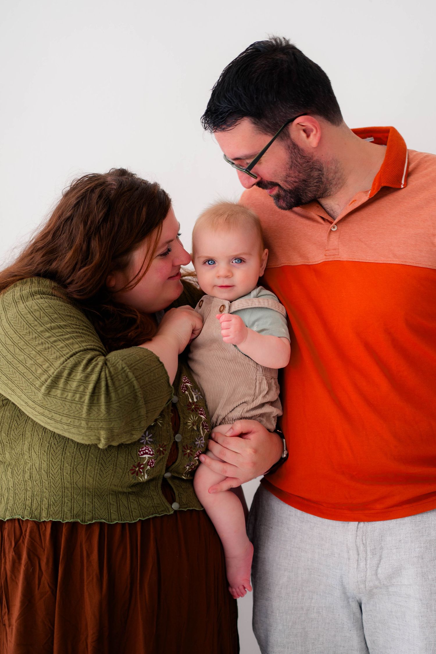 A family portrait of a couple lovingly holding their blue-eyed baby, the woman in green and the man in an orange polo shirt.