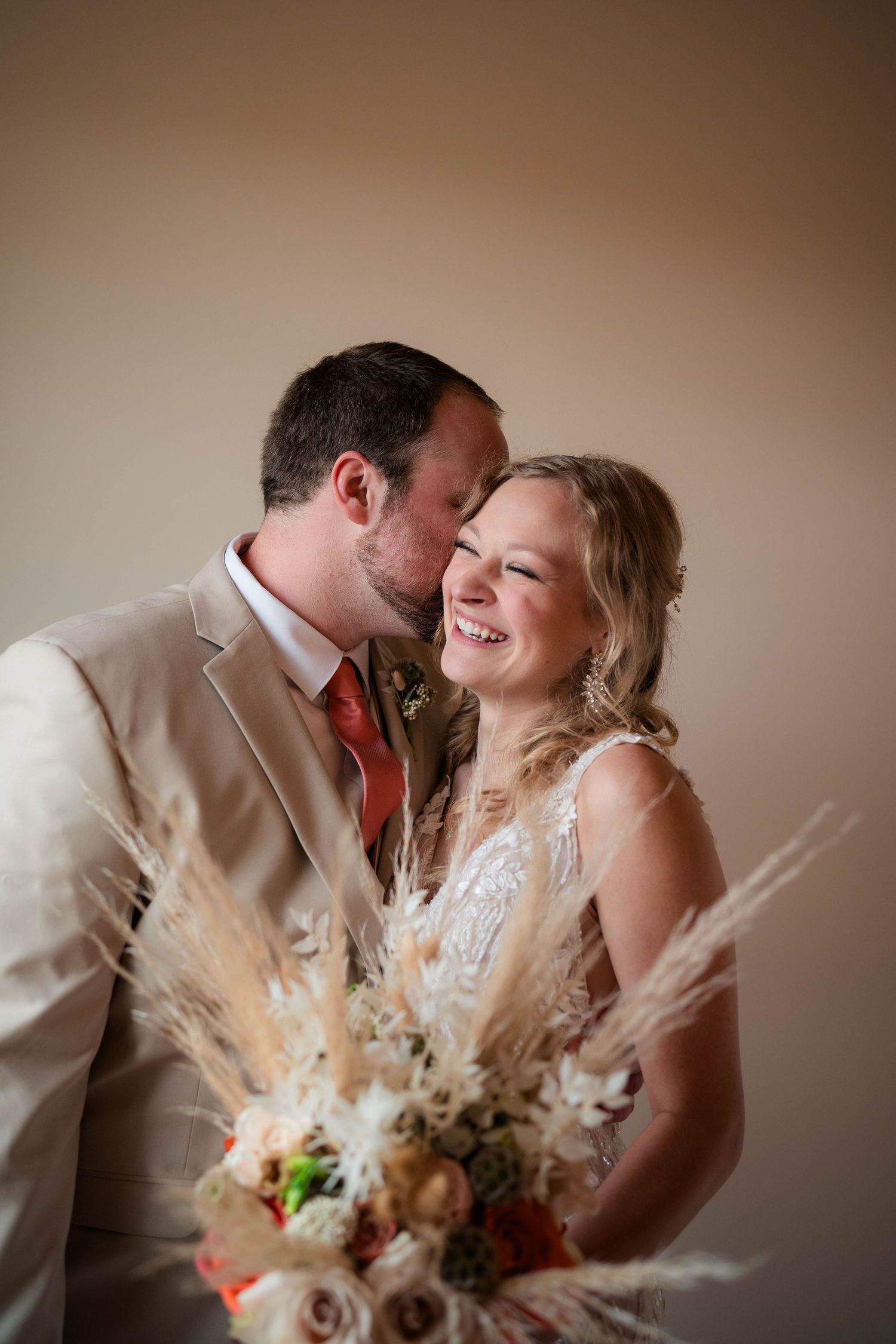 Smiling bride holds a boho pampas grass bouquet as groom kisses her cheek in a beige suit.