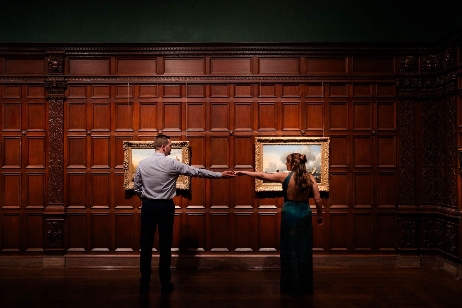 Couple holding hands while viewing paintings on wood-paneled wall in elegant museum setting.