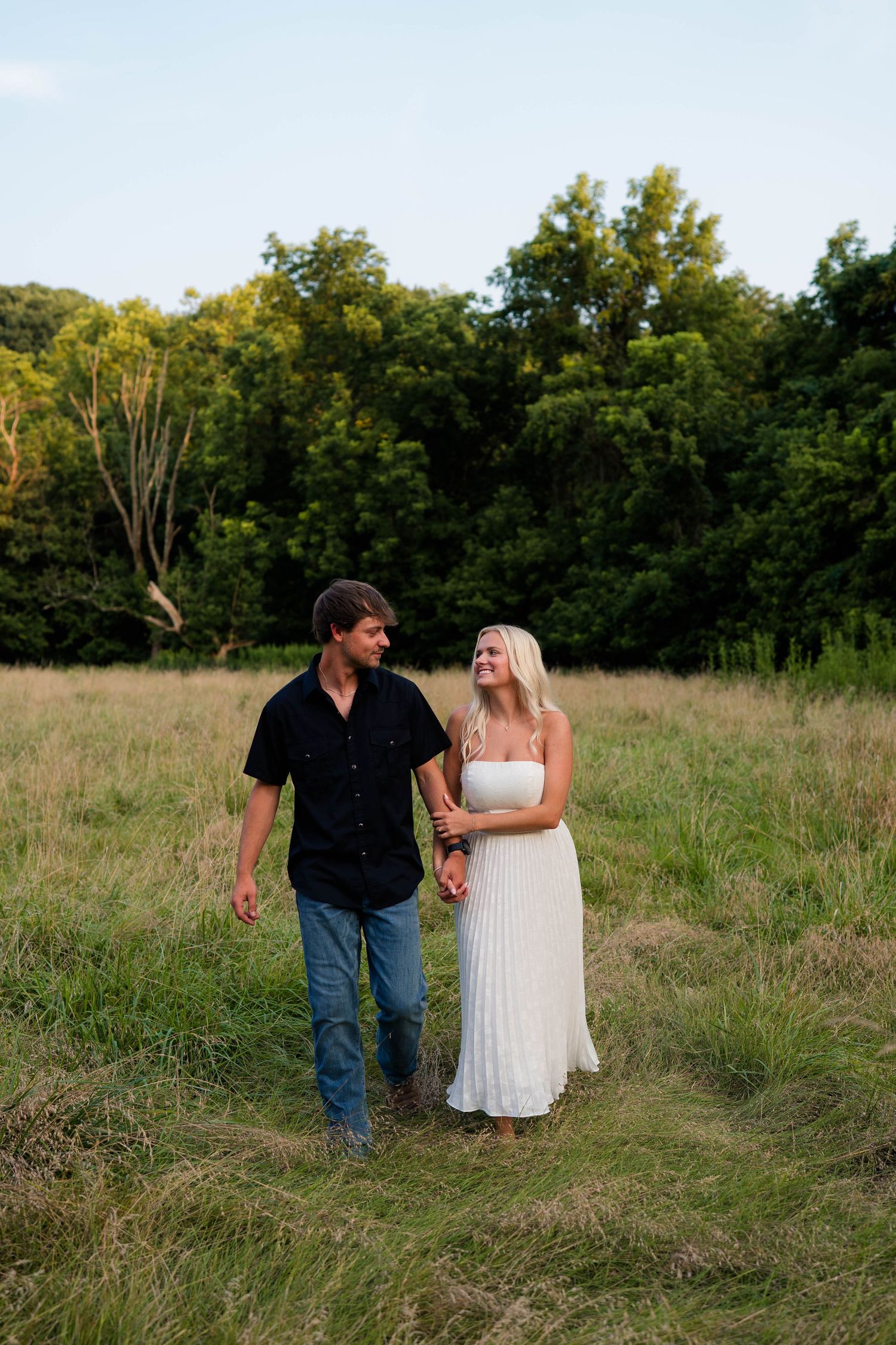A happy couple walks hand in hand through a lush green meadow, woman in white dress smiling at her partner.