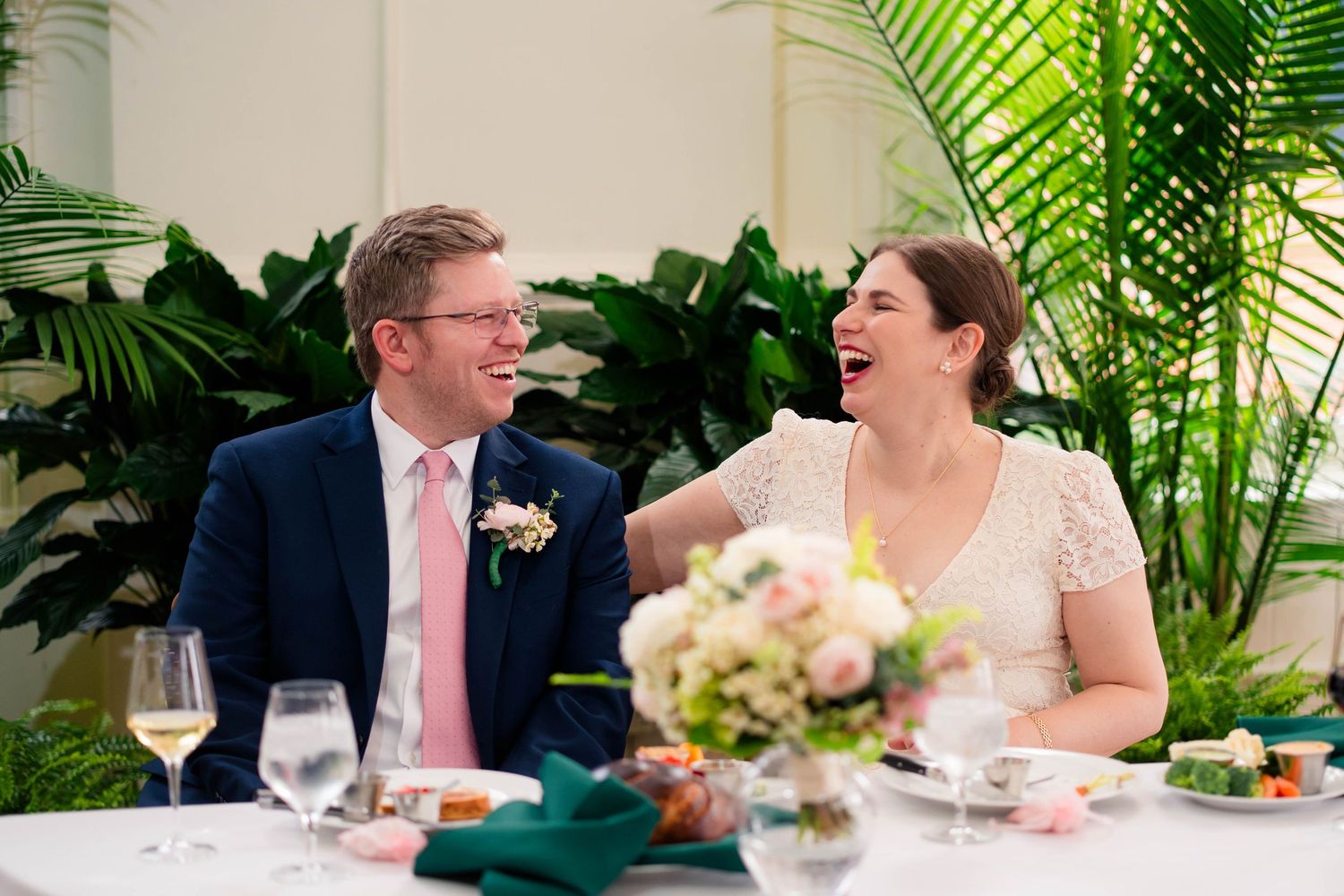 Laughing bride and groom share joyful moment at sweetheart table with floral centerpiece.