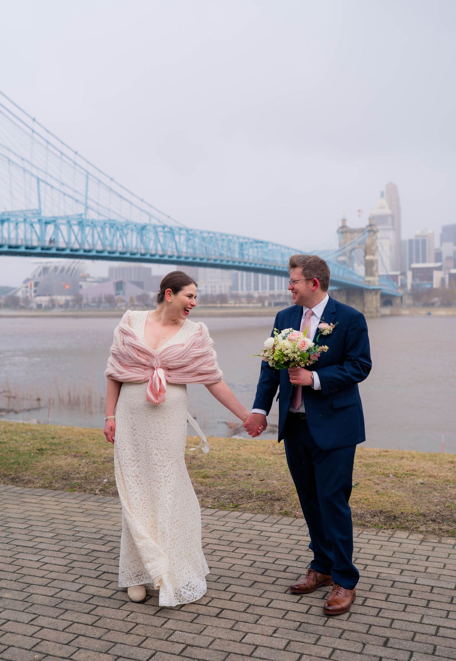 Laughing bride in lace gown and pink shawl holds hands with groom in navy suit by a river bridge.
