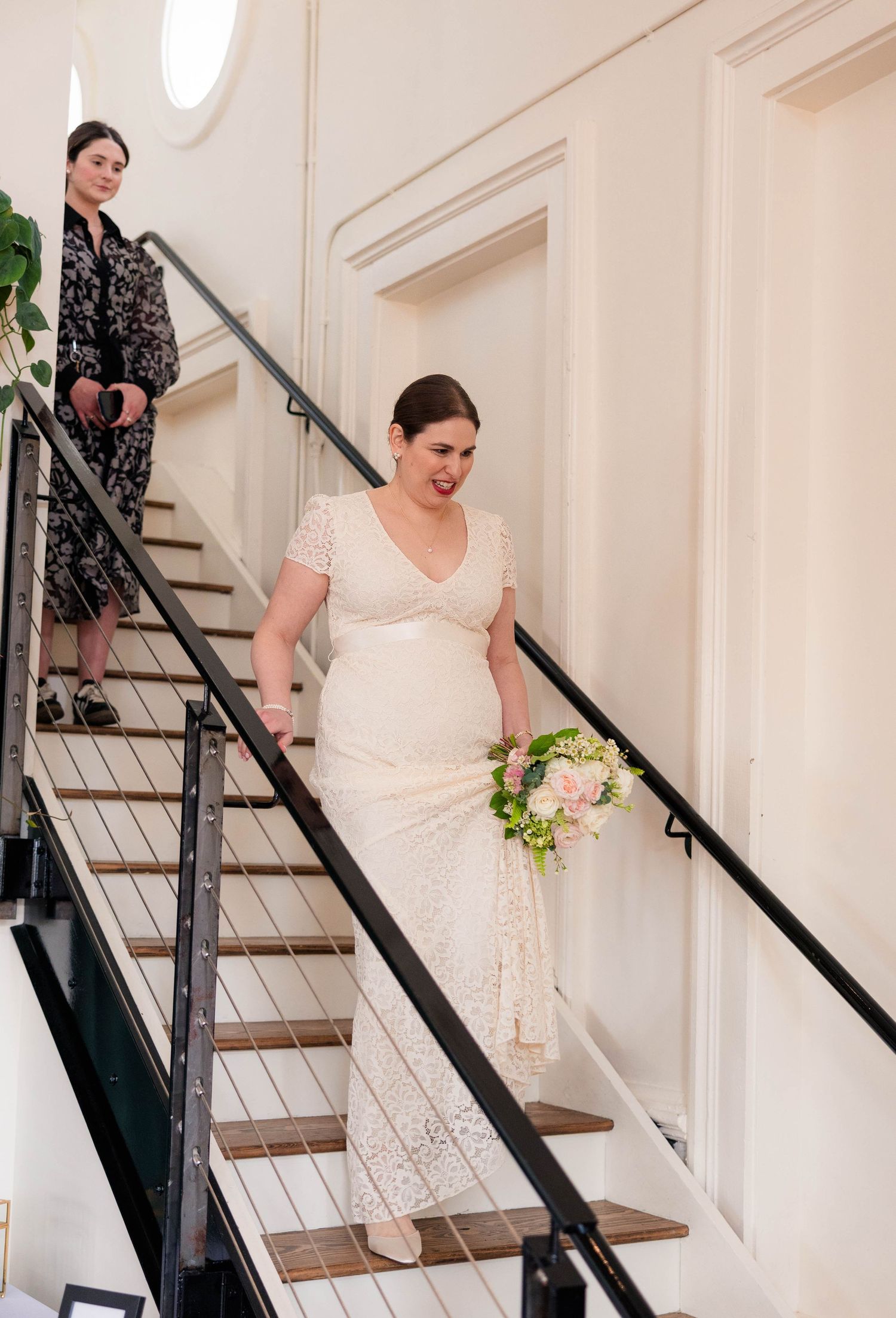 Bride in lace wedding gown carries pink bouquet while descending staircase at wedding venue.
