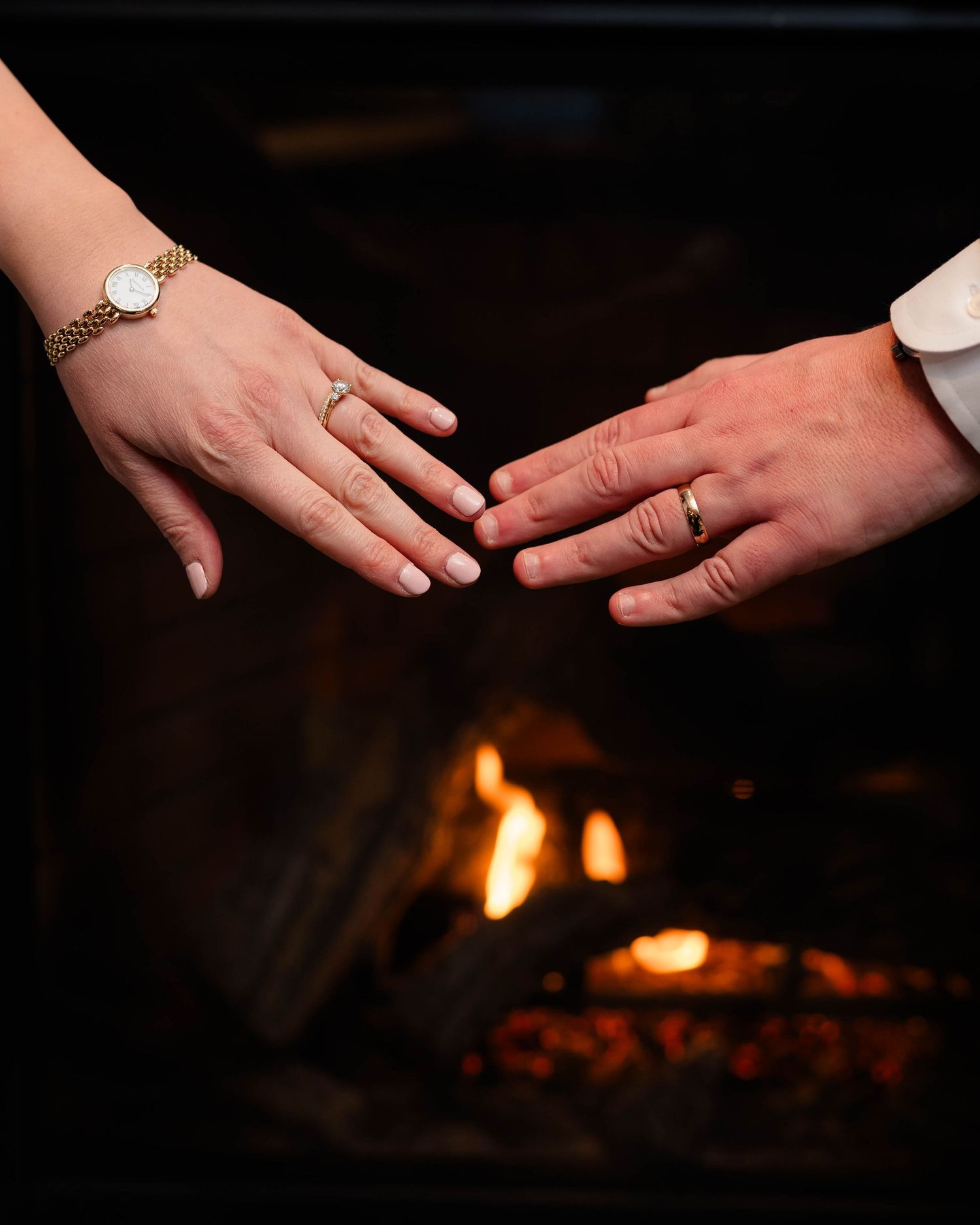 Bride and groom displaying wedding rings near a glowing fireplace, bride wearing a gold bracelet watch.
