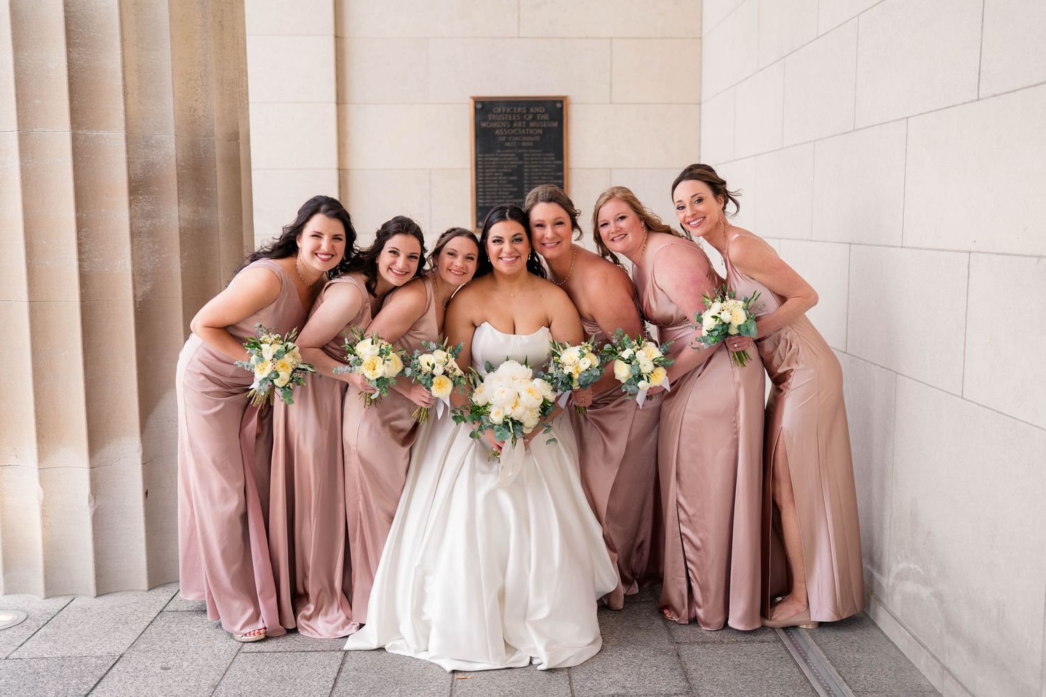 Bride poses with five bridesmaids in dusty rose gowns holding white bouquets against stone columns.