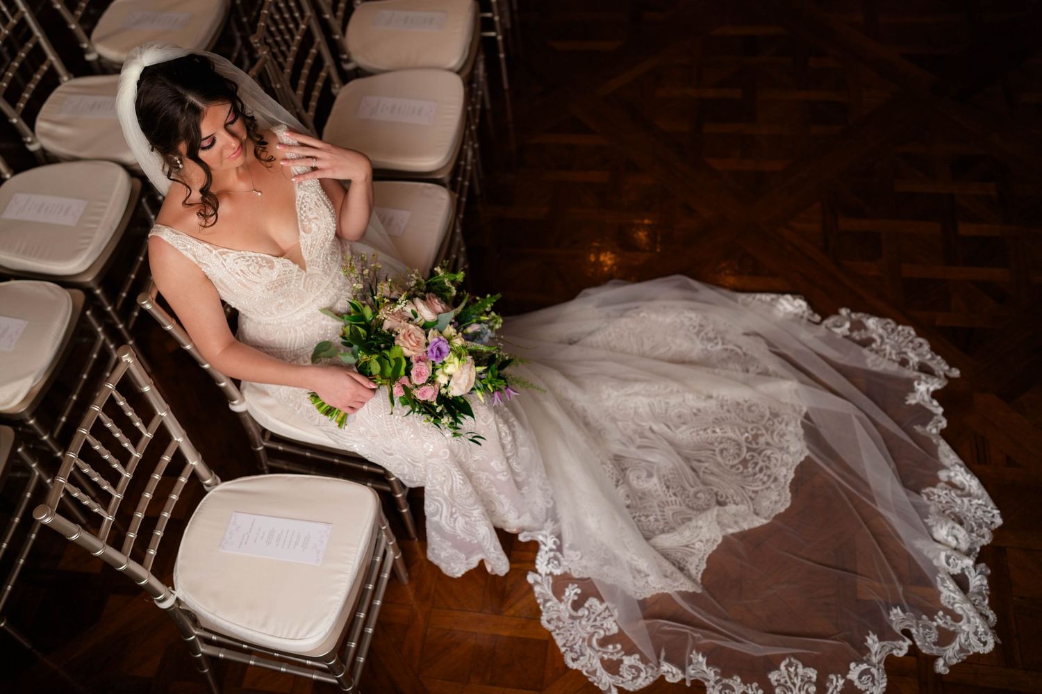 Bride in lace gown with long veil seated among chiavari chairs holding a floral bouquet.
