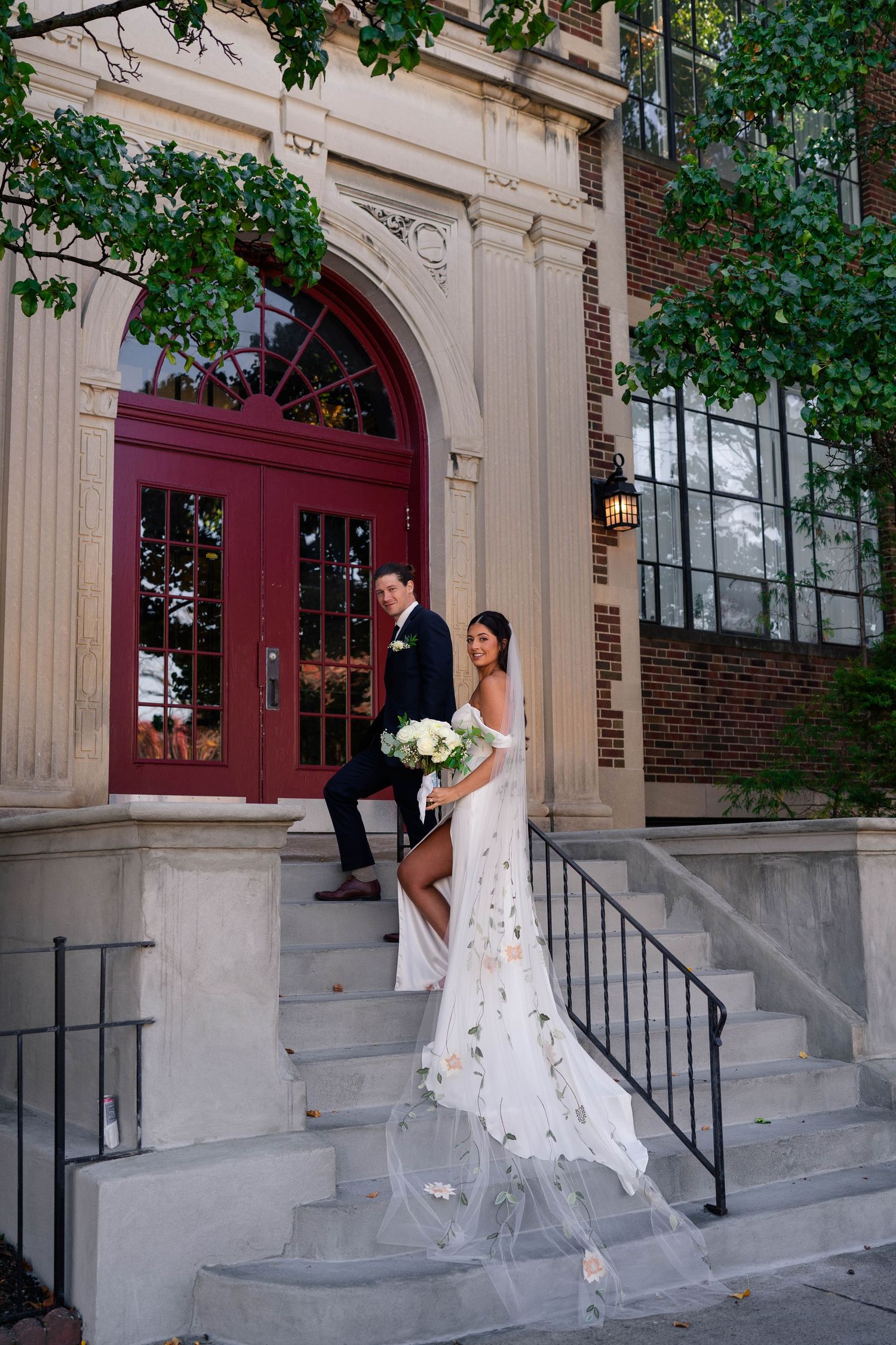 Bride and groom pose on ornate stone steps before elegant red arched doors of brick building.