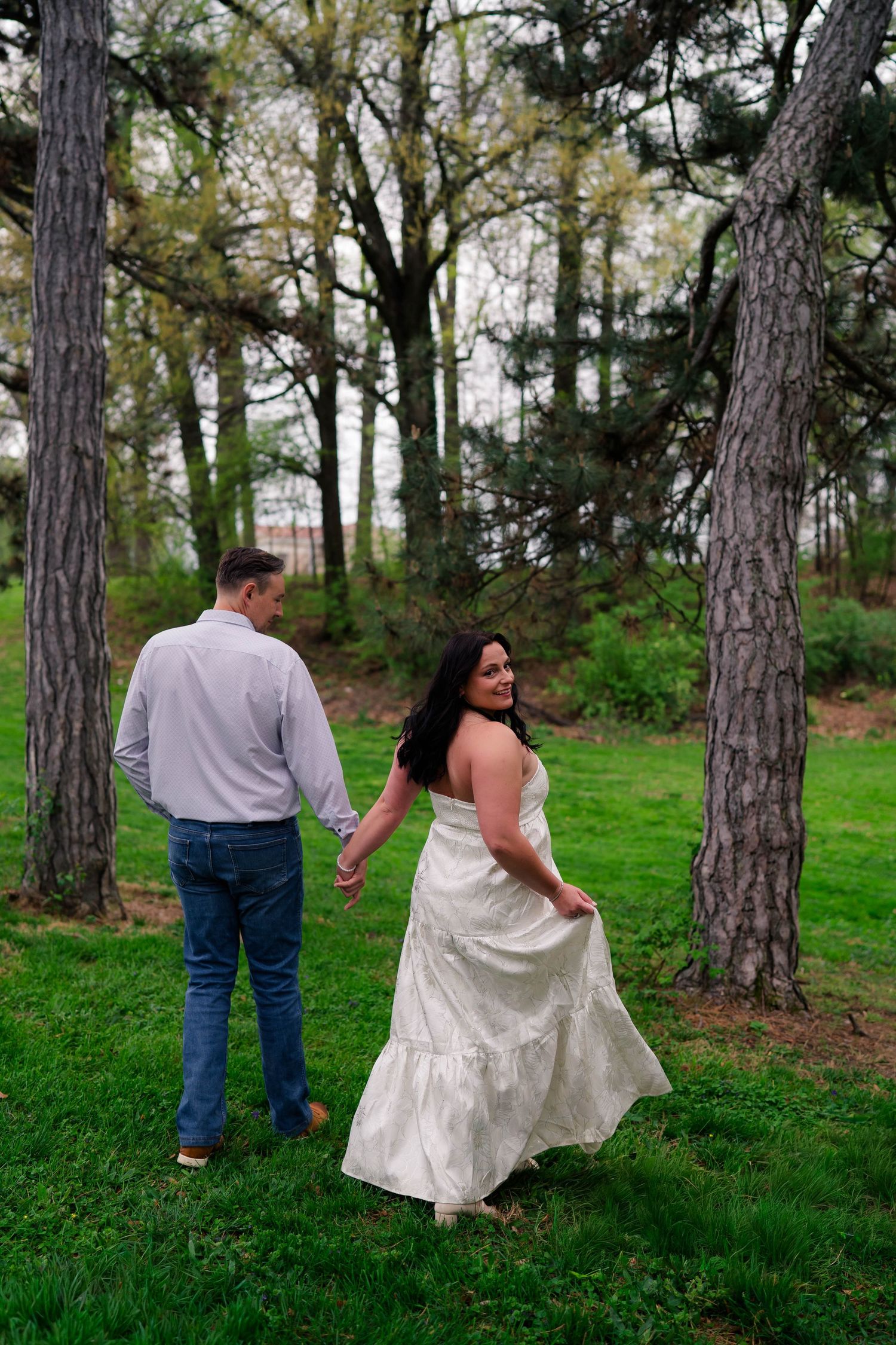 A couple walks hand-in-hand through a wooded park, the woman glancing back in a white dress.