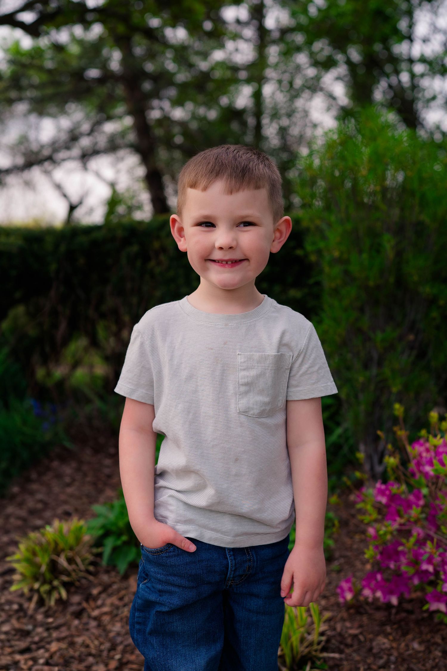 Young boy smiling in gray t-shirt and jeans standing in a lush garden with colorful flowers.