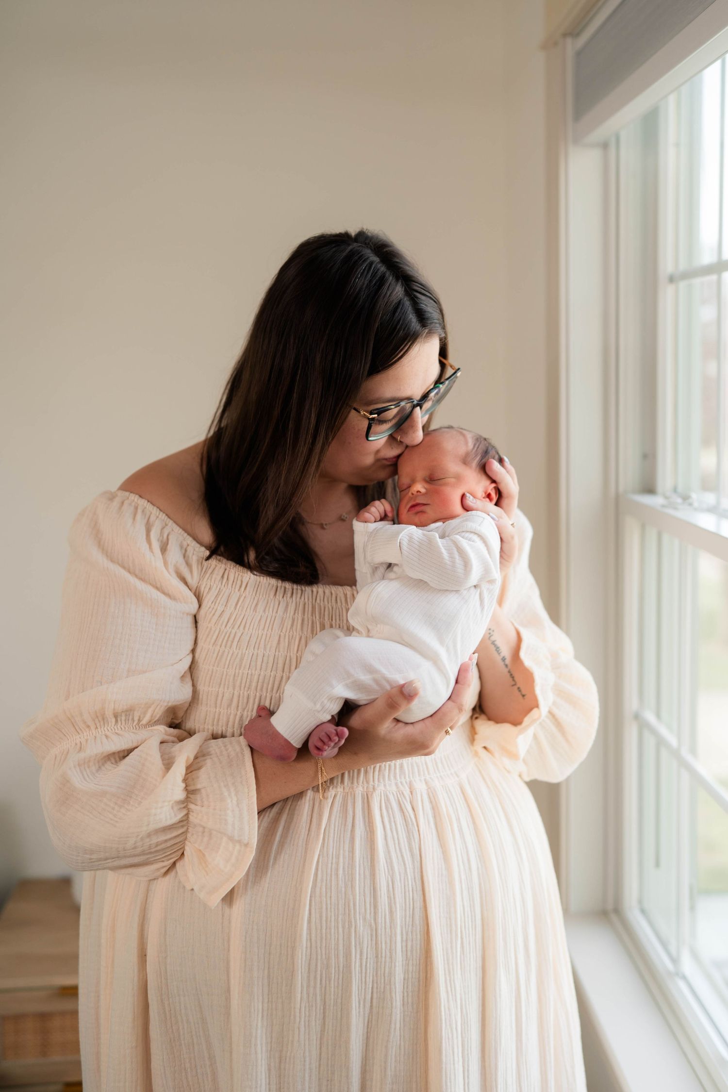 A mother in a cream dress tenderly kisses her newborn baby while standing near a bright window.