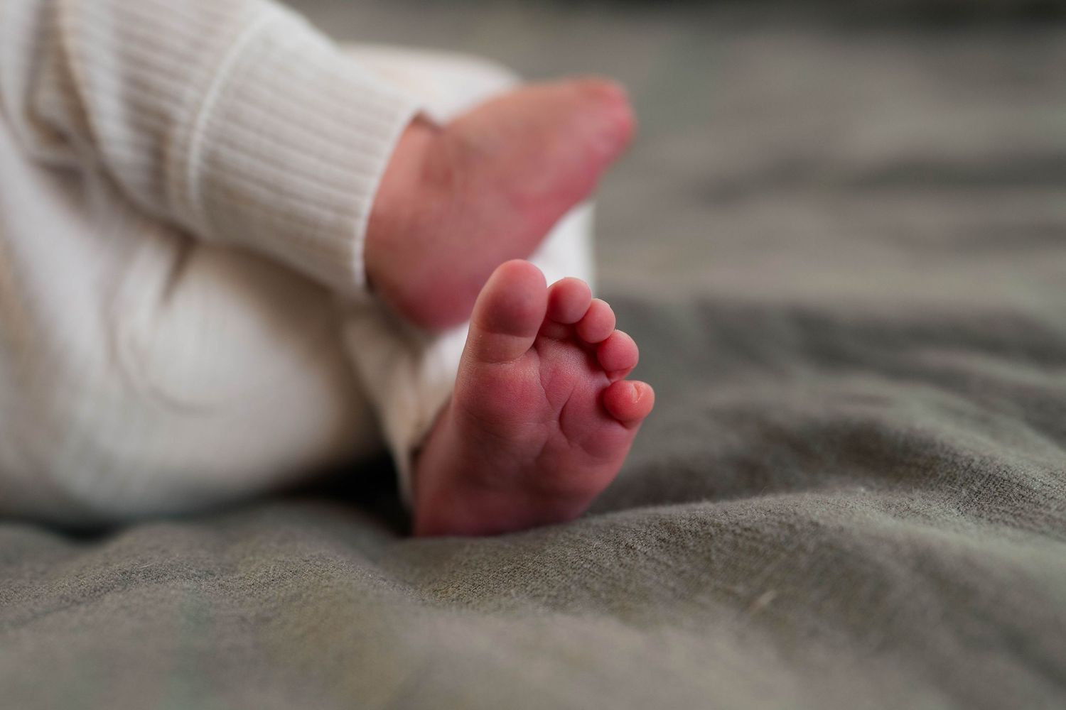 Newborn baby feet resting on gray blanket in white ribbed onesie.