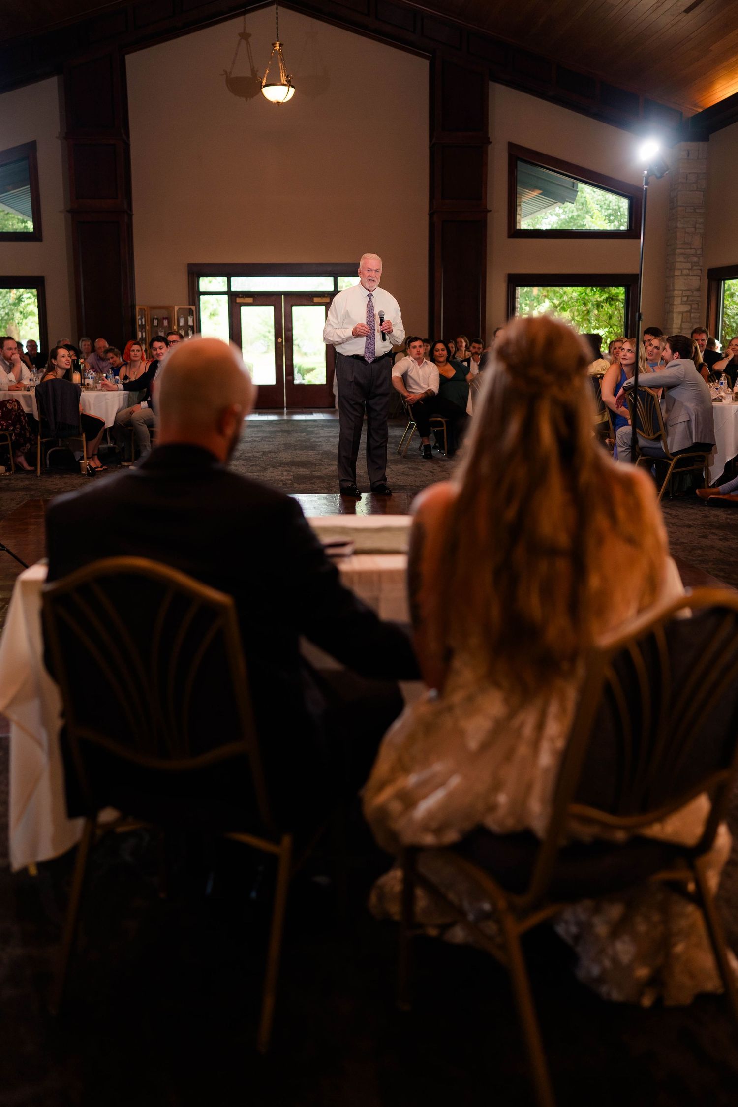 Wedding guest delivers heartfelt speech to bride and groom seated at sweetheart table in elegant reception hall.