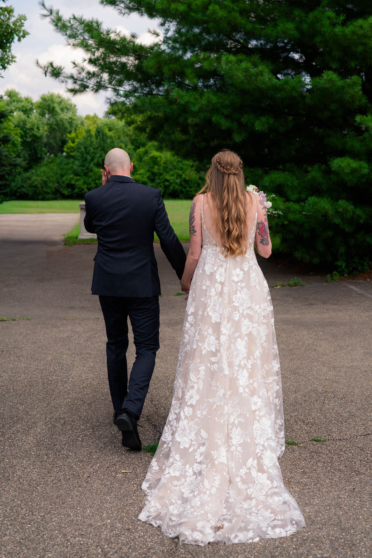 Newlyweds walk hand in hand away from the camera through a lush green park after their ceremony.