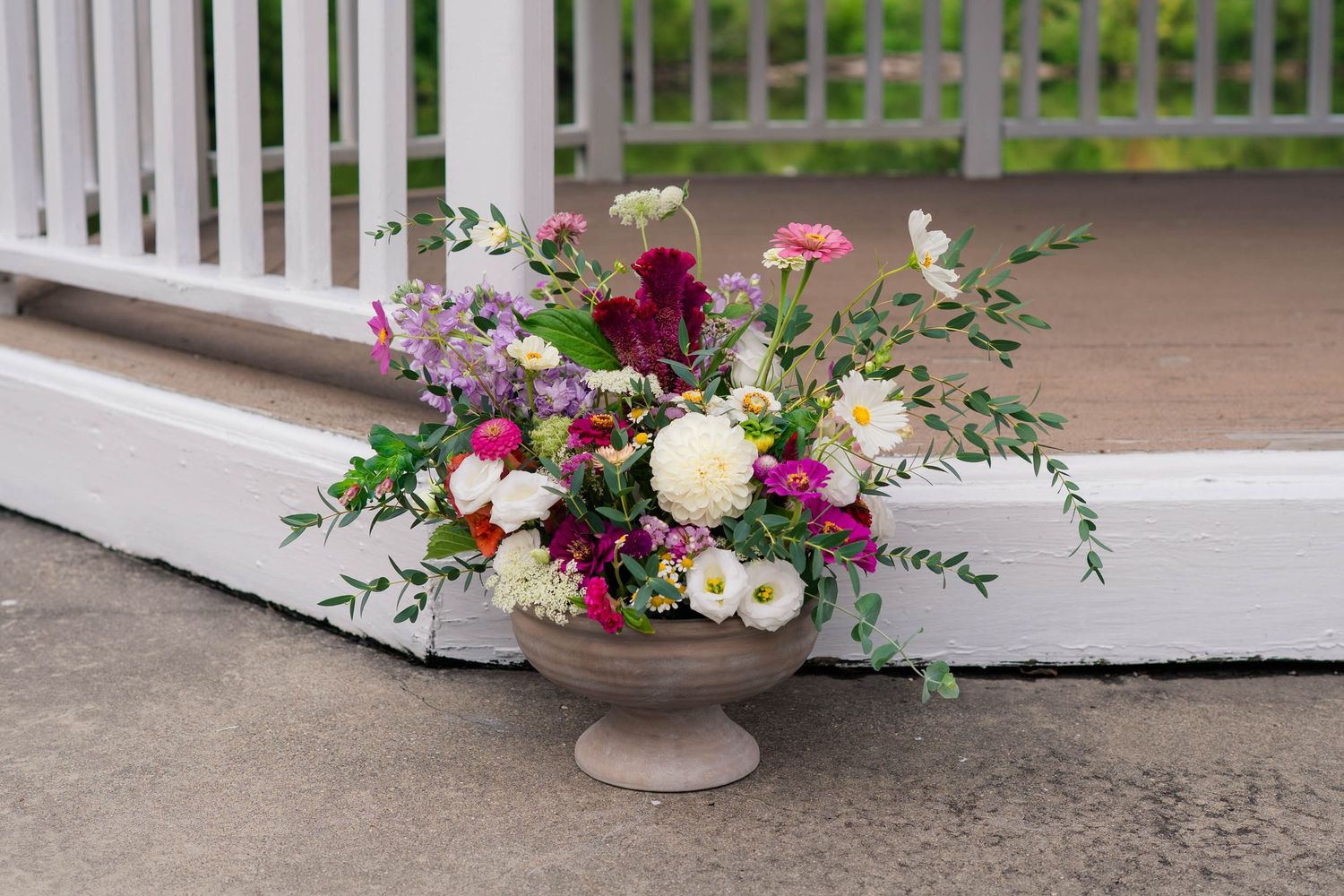 Lush garden floral arrangement in stone urn featuring pink, white, and burgundy blooms with greenery at wedding venue.