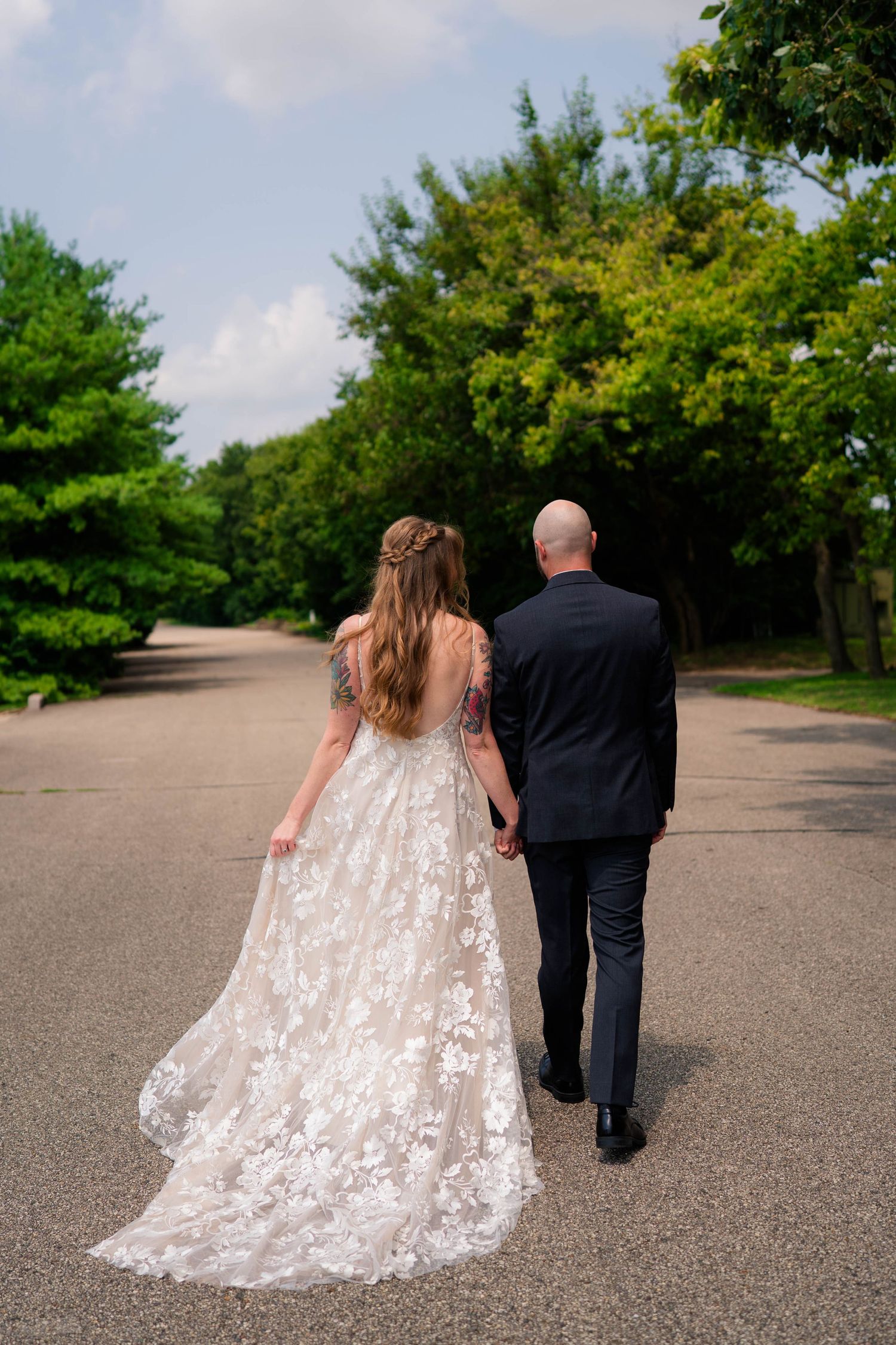 A bride and groom walk hand in hand down a tree-lined path, showing the bride's lace gown train.