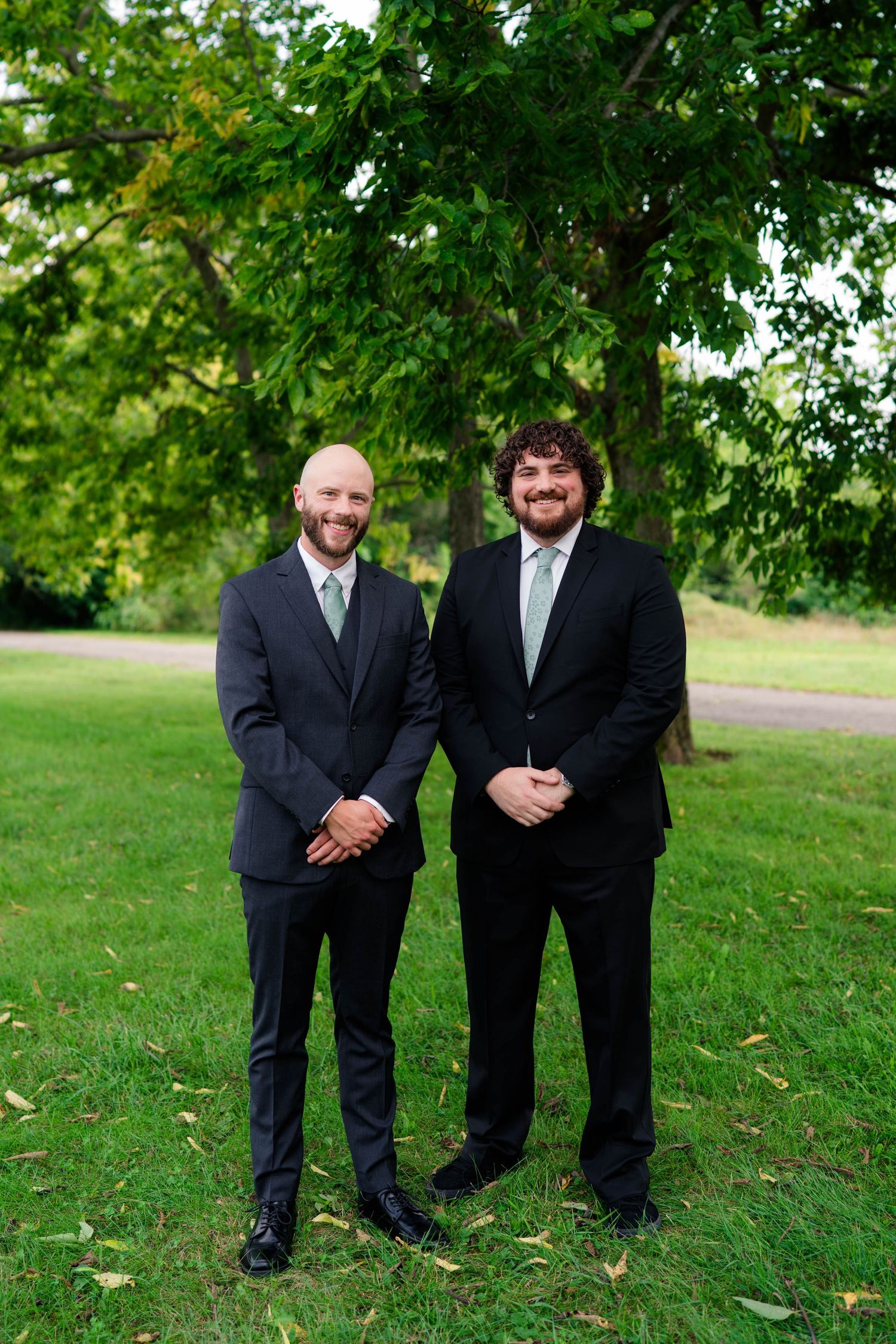 Two groomsmen in dark suits with sage green ties smile outdoors on green grass with trees behind them.