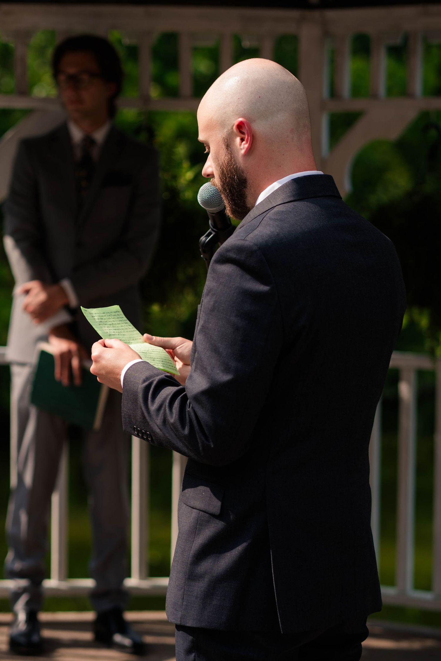 Bald groom reads vows from a green card at an outdoor wedding ceremony at a white gazebo.