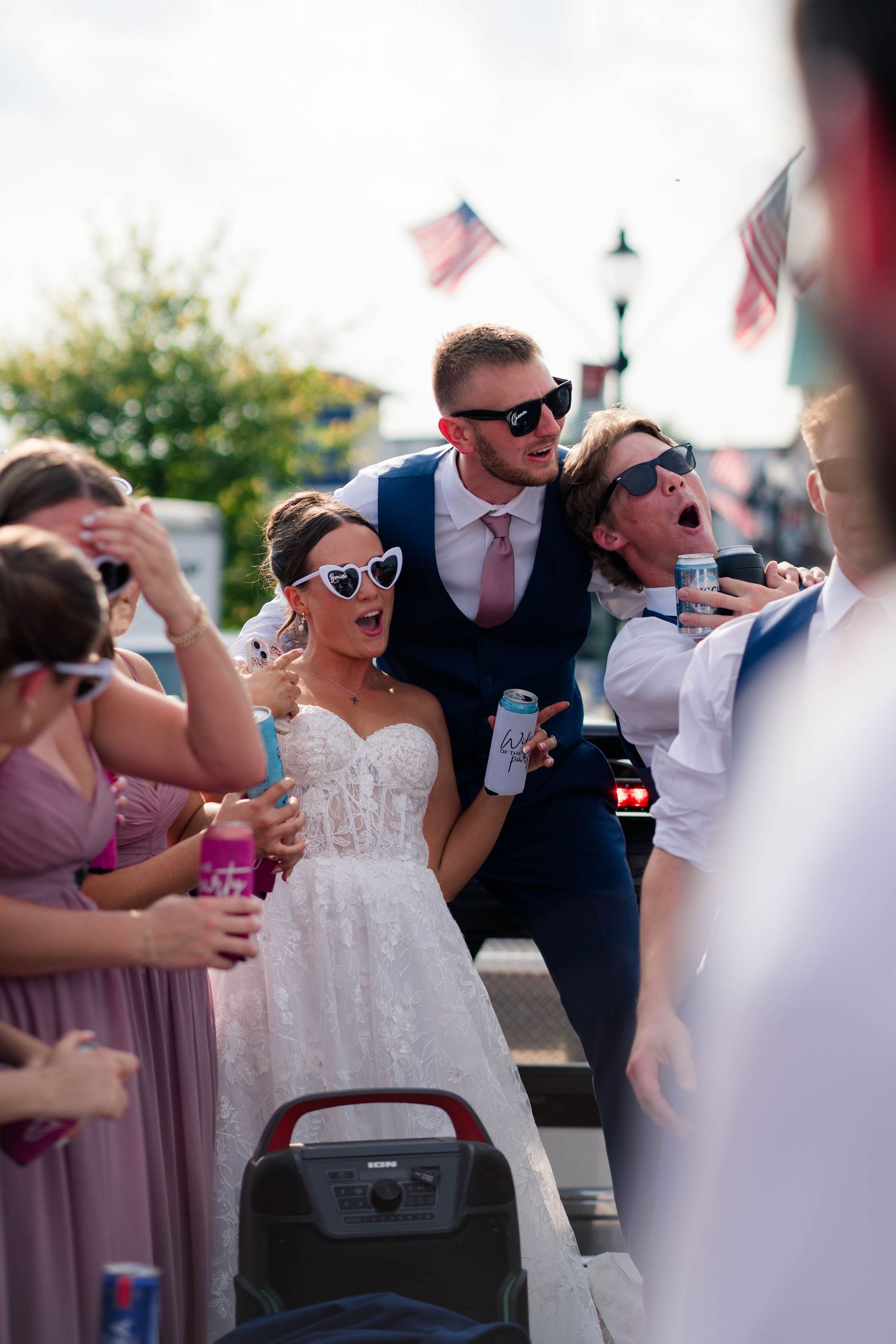 Bride and groom celebrate with wedding party on truck bed wearing sunglasses at fun outdoor reception.