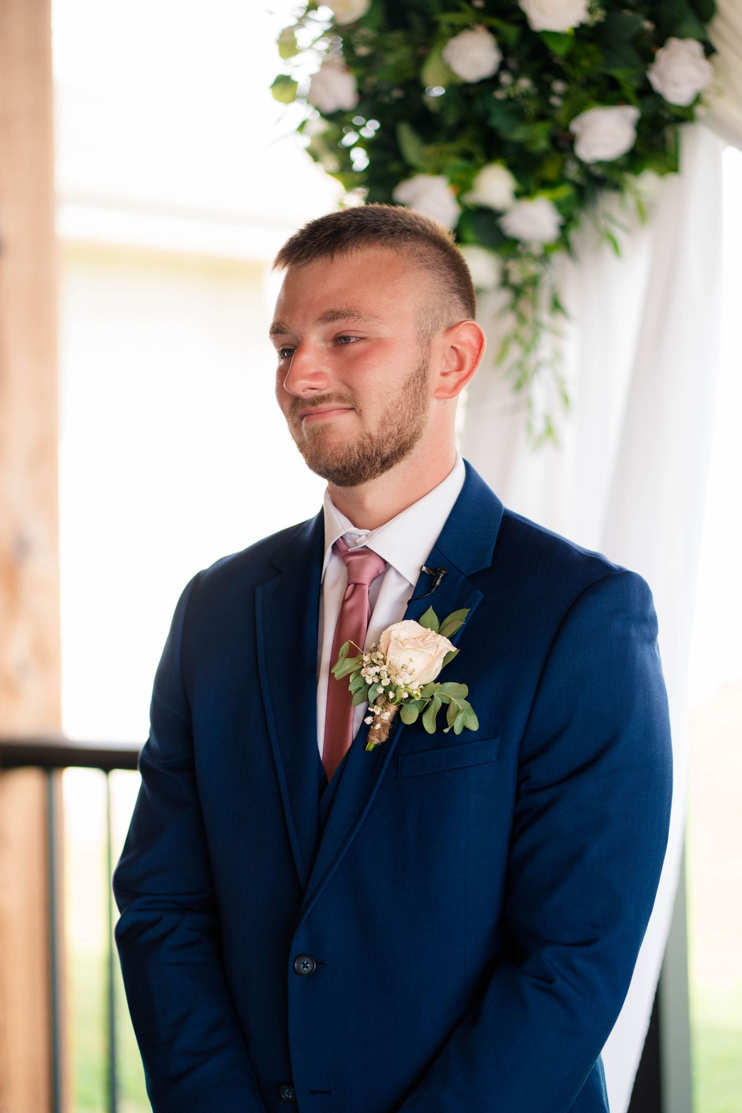 Groom in navy suit with blush boutonniere smiling emotionally at wedding ceremony with floral arch backdrop.