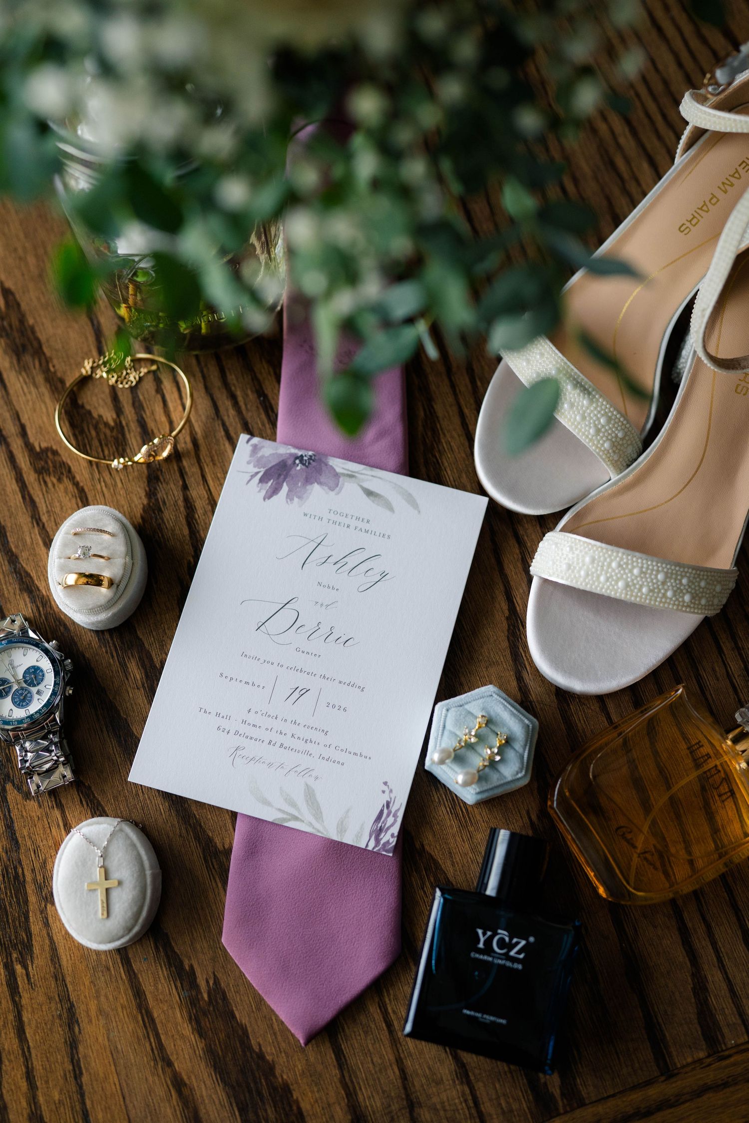 Wedding flat lay with invitation, purple tie, heels, rings, watch, cross pendant, cologne, and floral accents on wood.