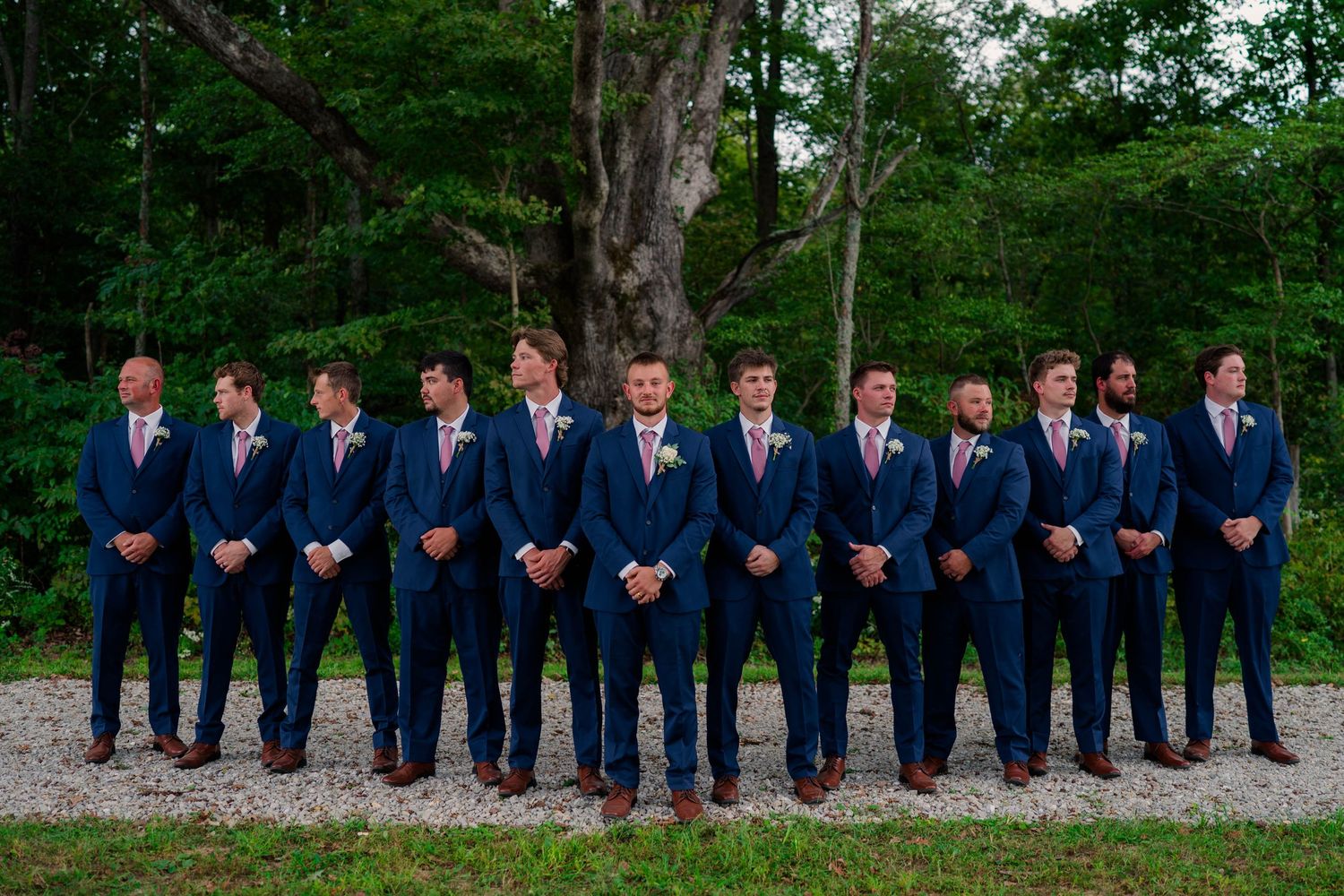 Wedding party groomsmen lineup in matching navy suits with pink ties on gravel path outdoors.