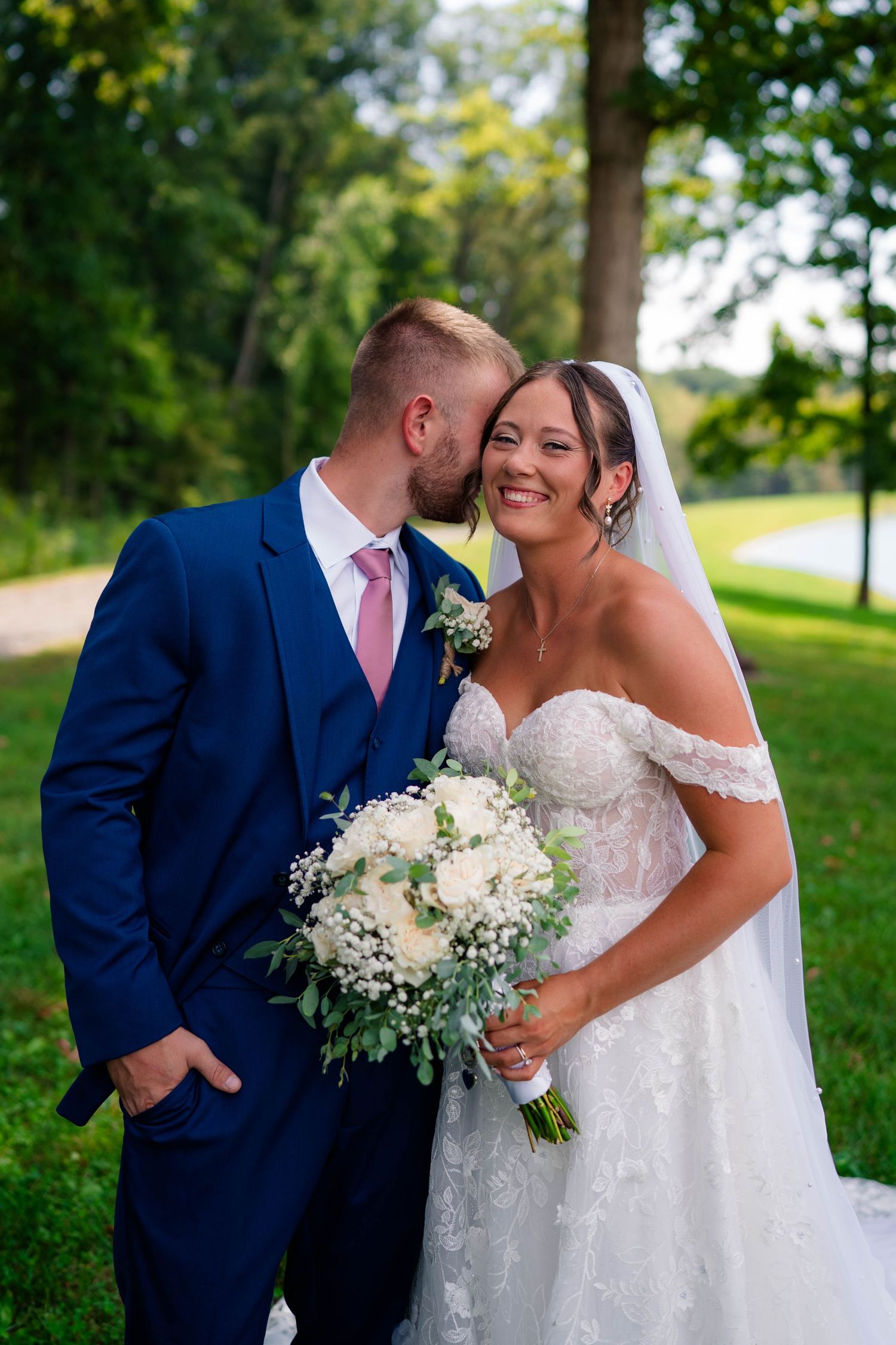 Smiling bride holds white bouquet as groom in blue suit kisses her cheek outdoors.