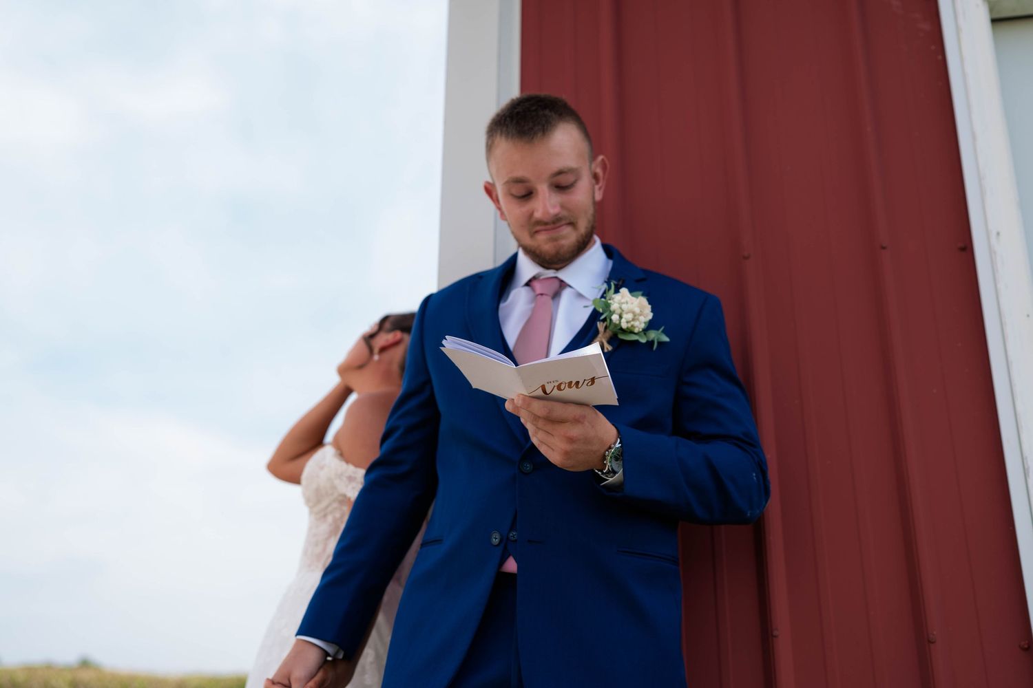 Groom in navy suit reading vows card leaning against red barn during first look with bride.