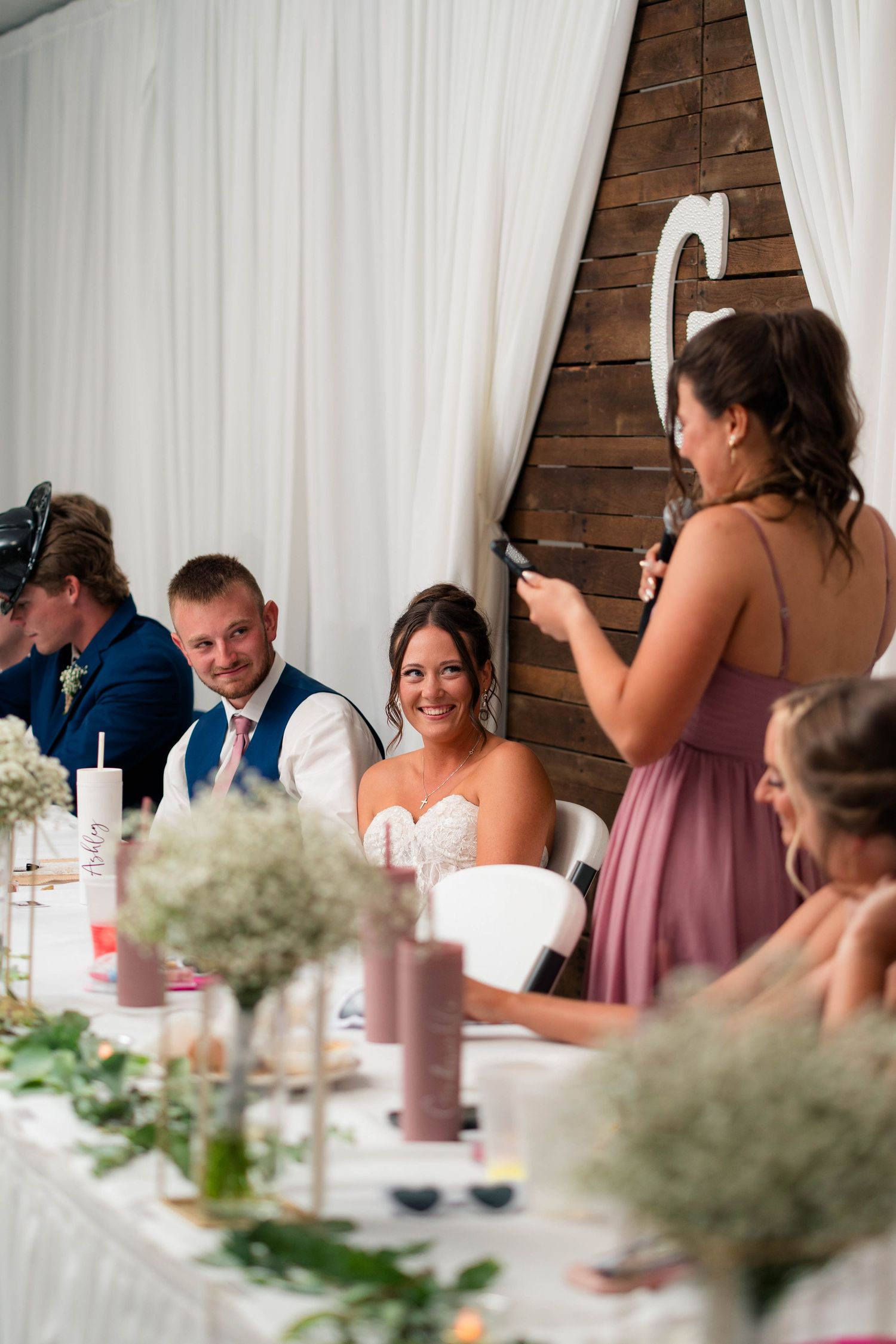 Bridesmaid in dusty rose dress gives touching microphone speech to beaming bride and groom at reception table.