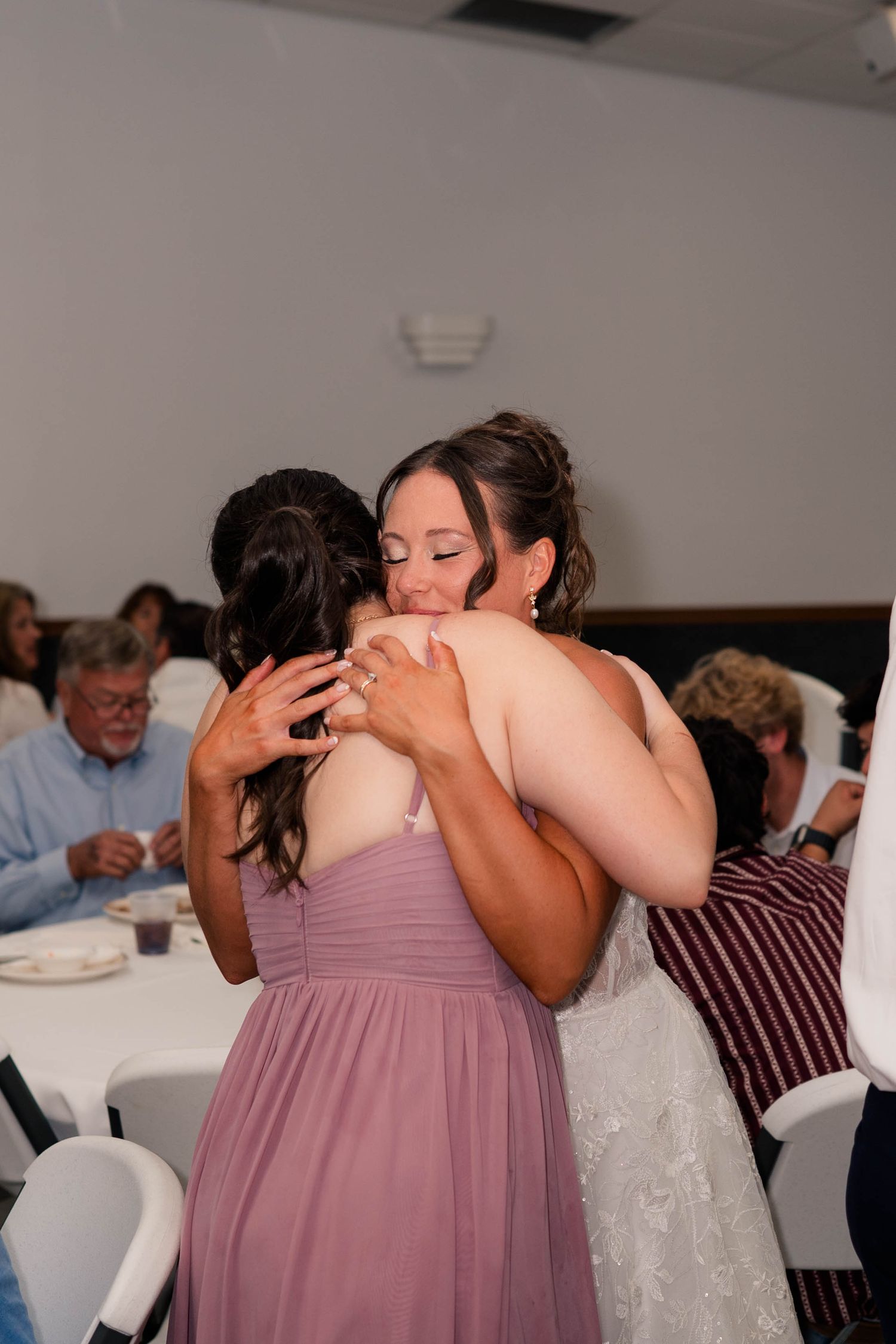 Bride in white lace gown shares an emotional hug with bridesmaid in dusty rose dress at wedding reception.