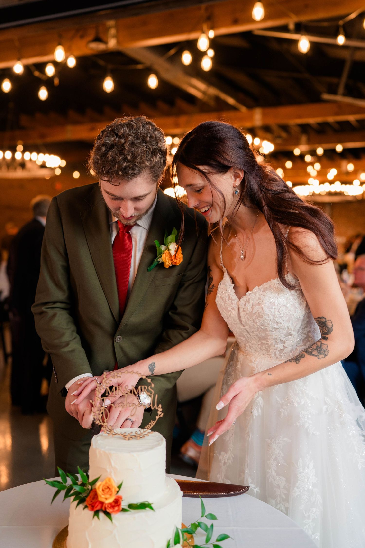 Bride and groom cutting their white wedding cake decorated with orange flowers under warm string lights.