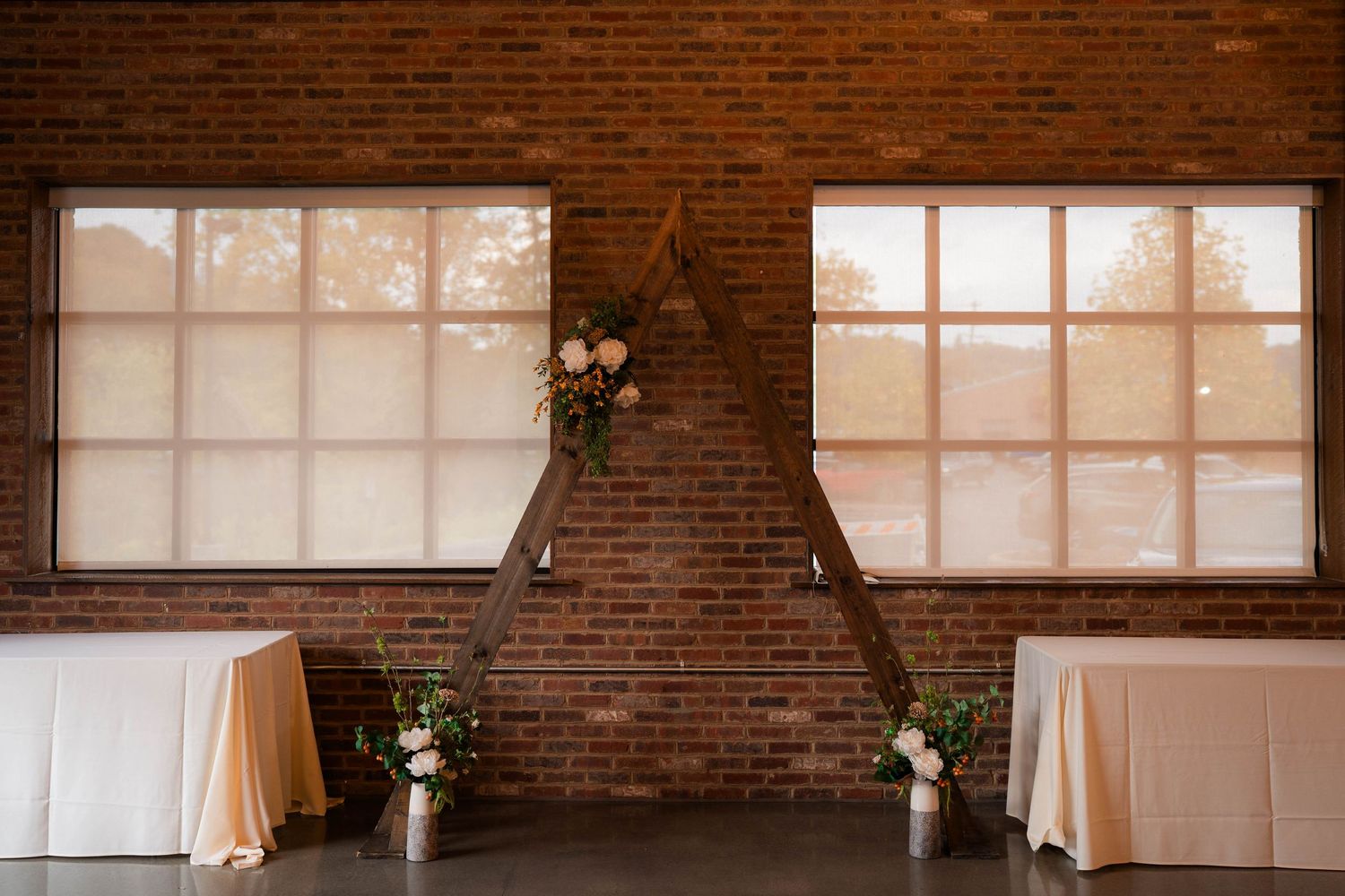 Rustic wedding ceremony arch with floral decor against exposed brick wall and large windows.