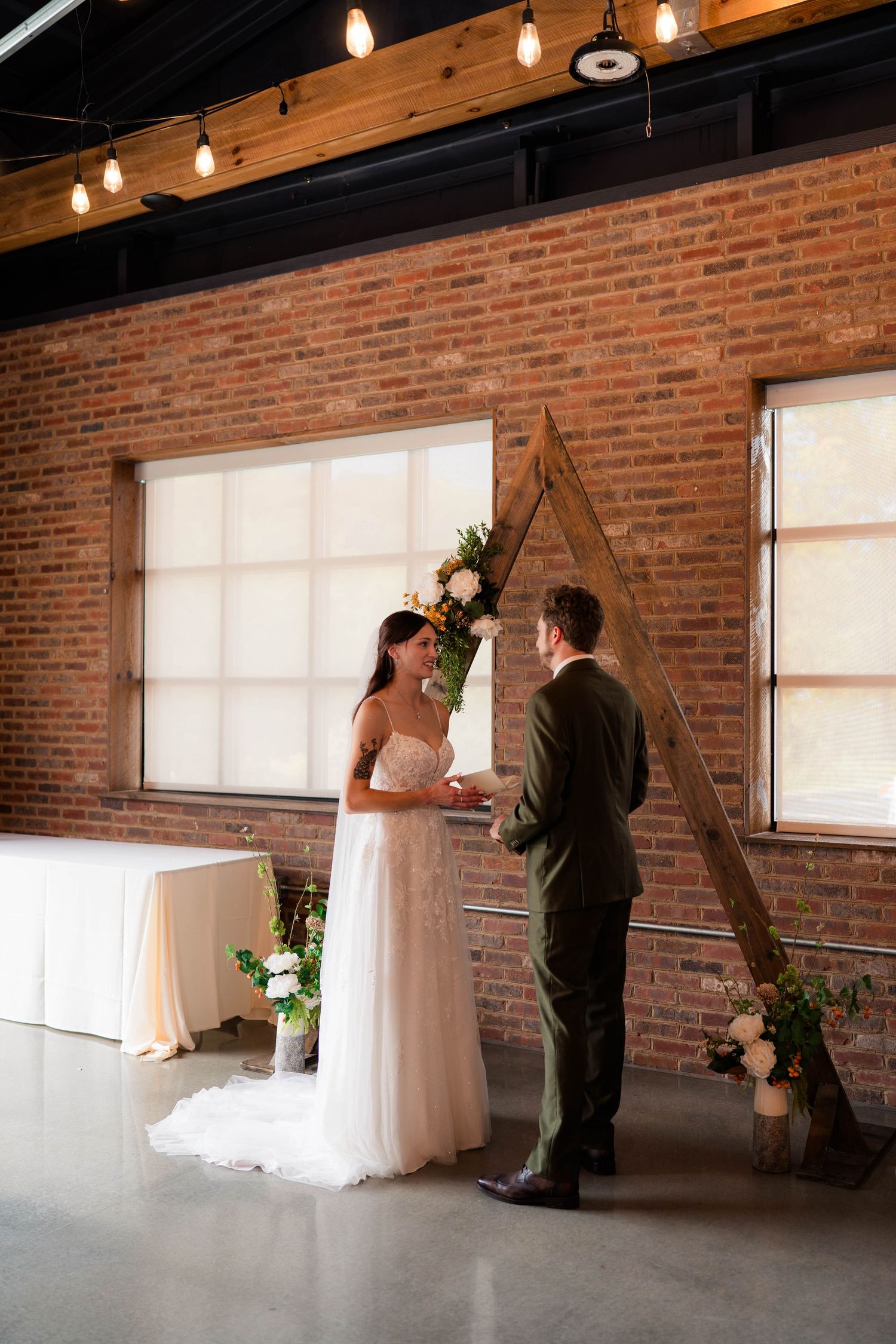 Bride and groom exchange vows at a rustic indoor wedding ceremony with exposed brick walls and string lights.