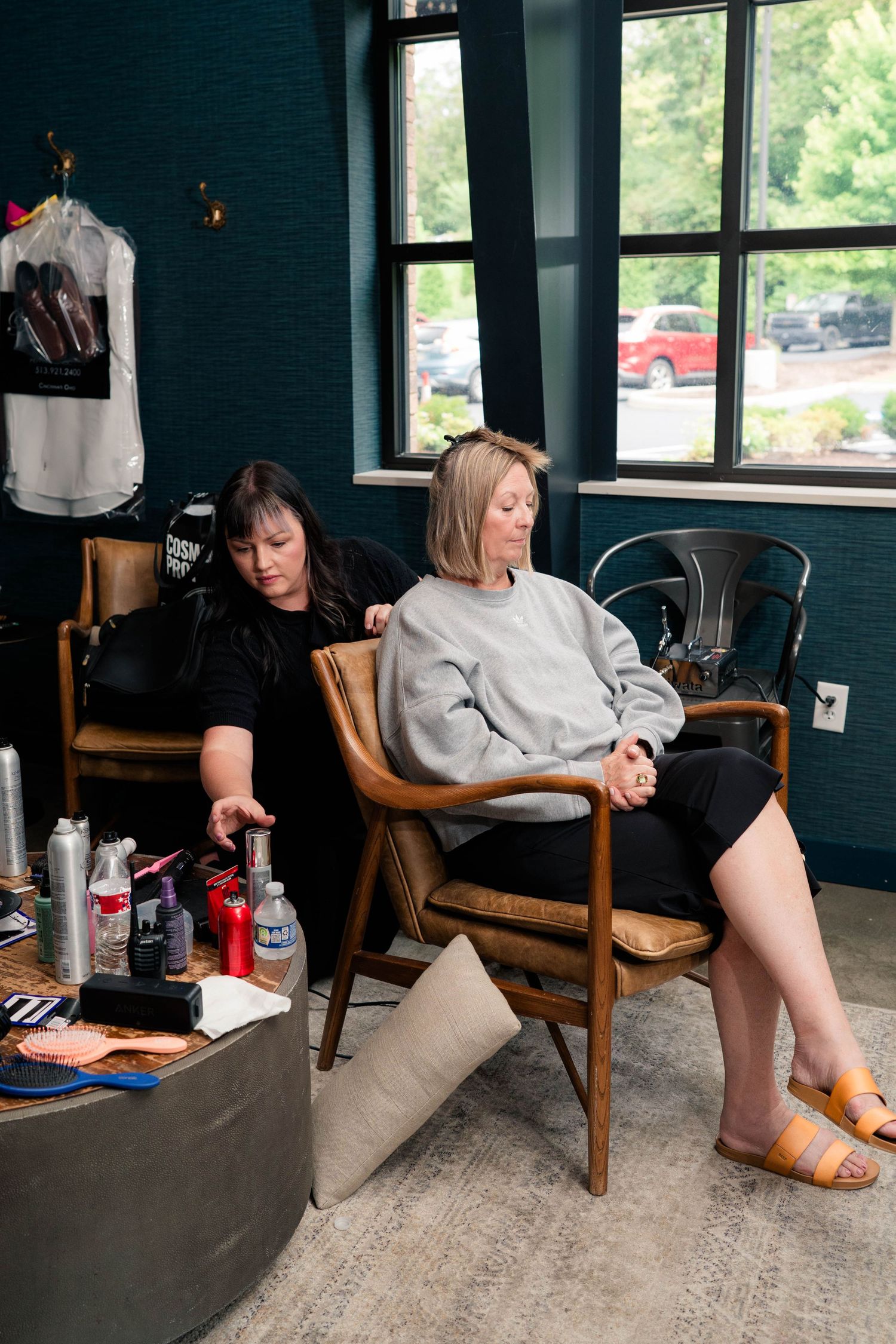 Hair stylist applying treatment to blonde client seated in wooden chair at modern teal-walled salon.