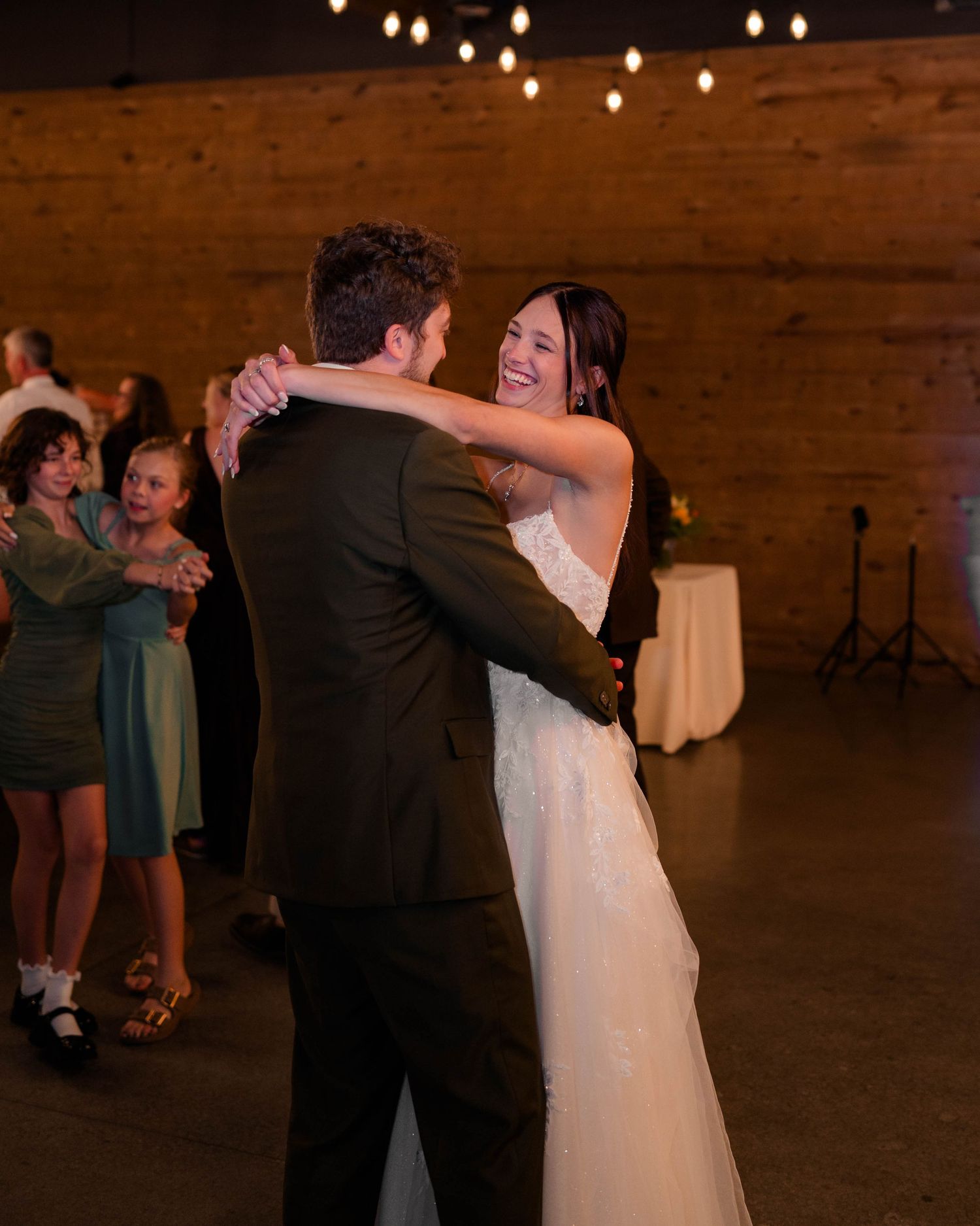 Radiant bride laughs joyfully while dancing with groom at rustic wedding reception.