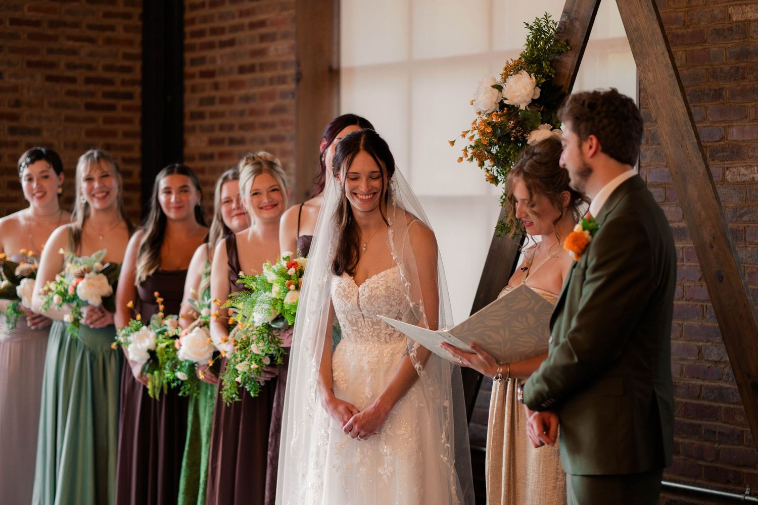 Bride smiles during vows surrounded by bridesmaids in sage green dresses at rustic brick venue.