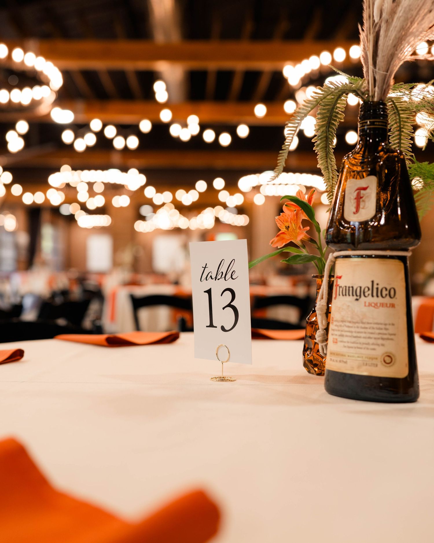 Table 13 centerpiece featuring Frangelico bottle with feathers and orange flowers at wedding reception.