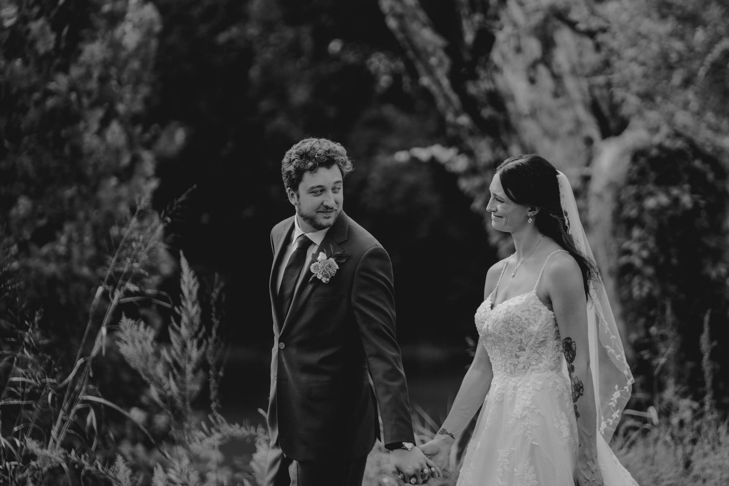 Black and white photo of bride and groom holding hands walking through lush garden on wedding day.