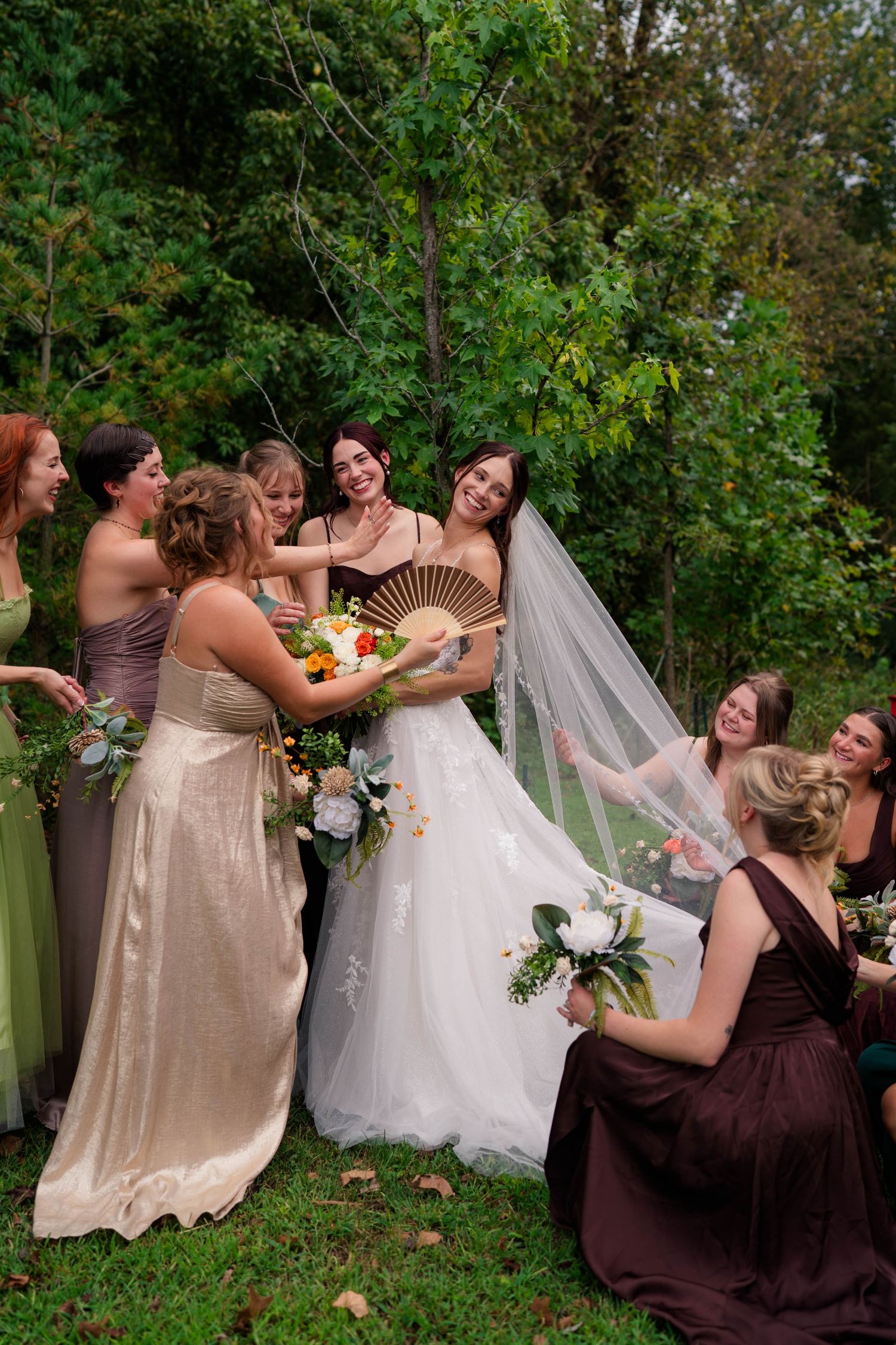 Bridesmaids in mixed earth-tone gowns surround laughing bride holding bouquet and fan on grassy outdoor setting.