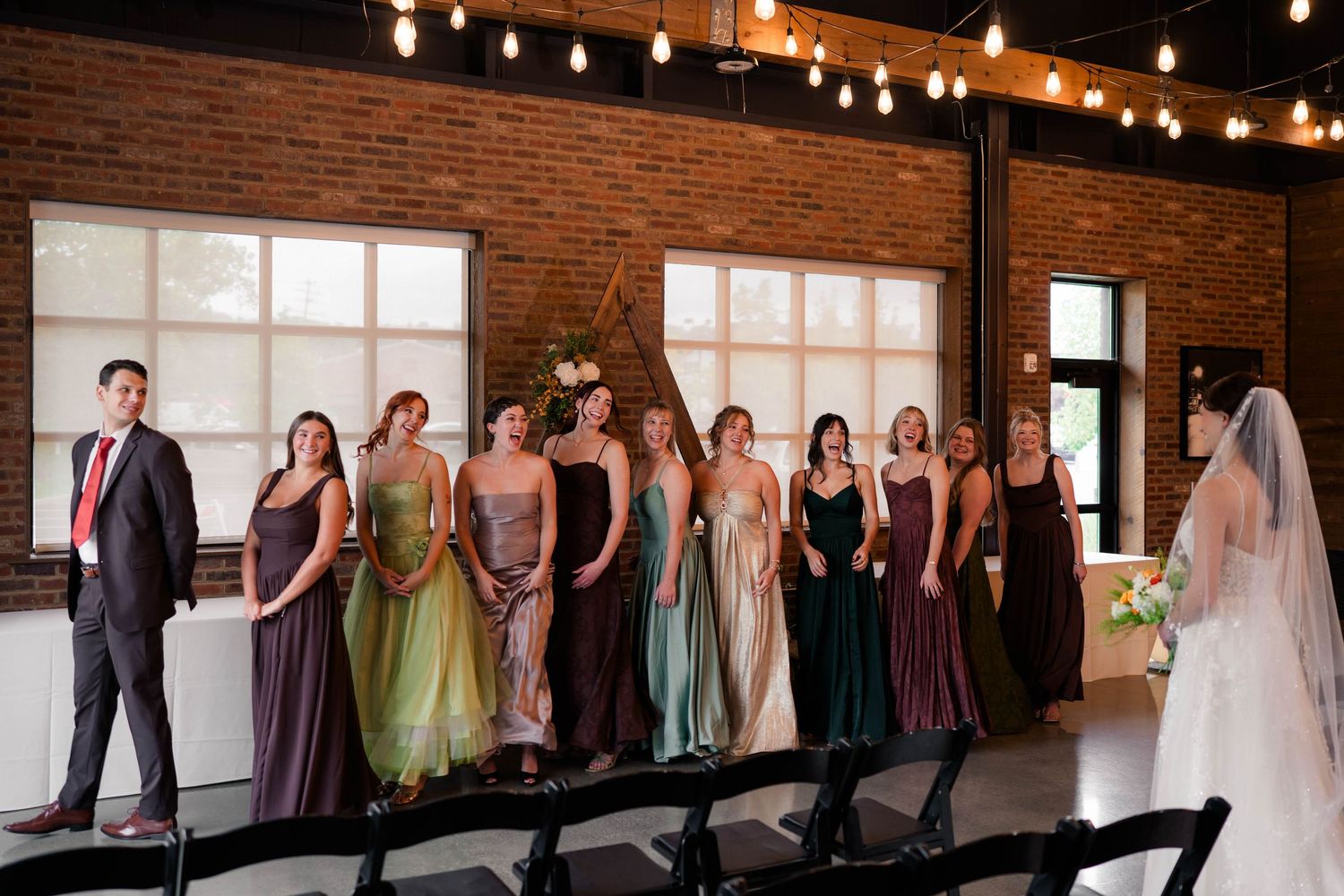 Wedding party lineup of bridesmaids in earth-tone dresses with groom in red tie at venue.