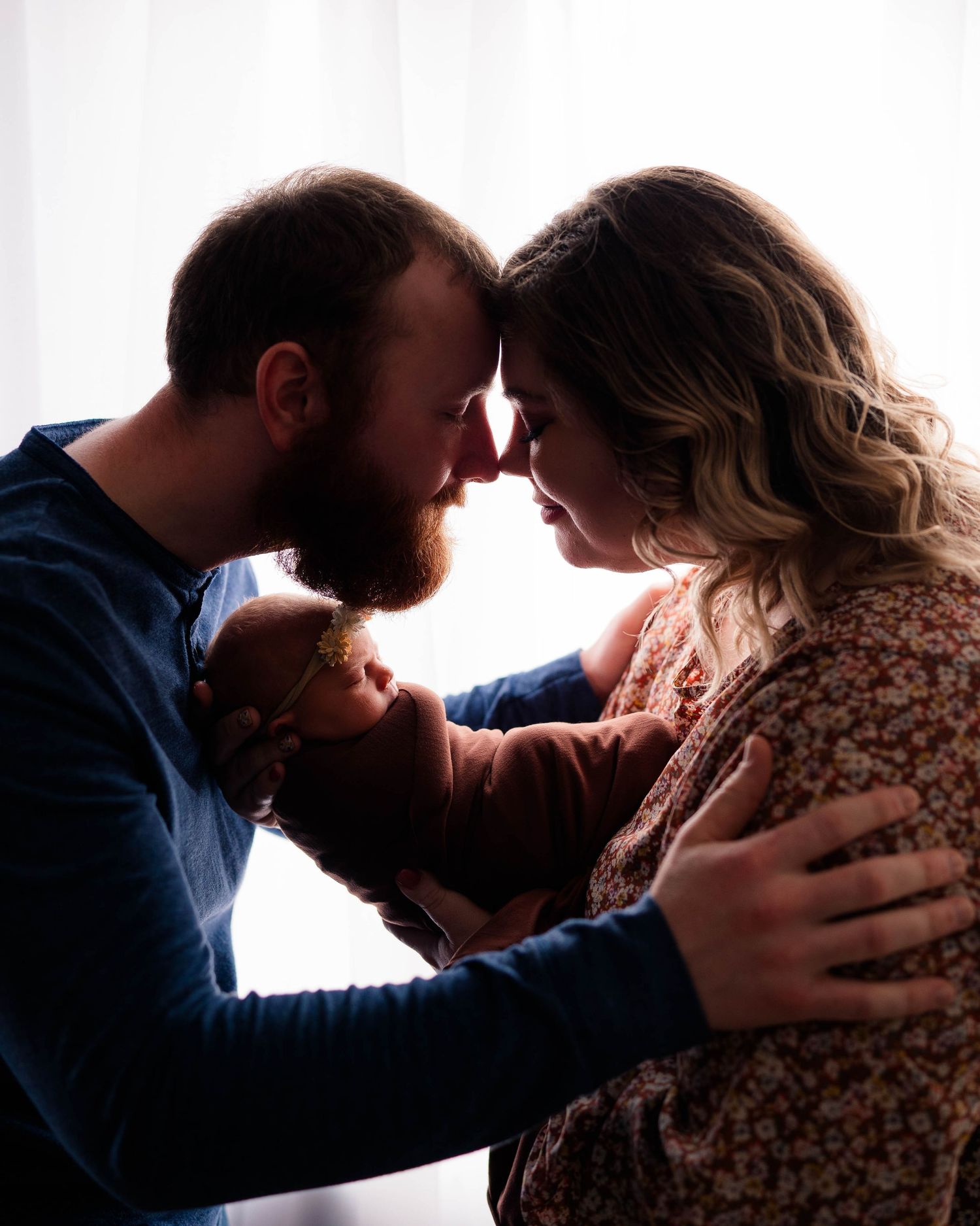 Silhouetted couple touching foreheads while lovingly holding their newborn baby near a bright window.