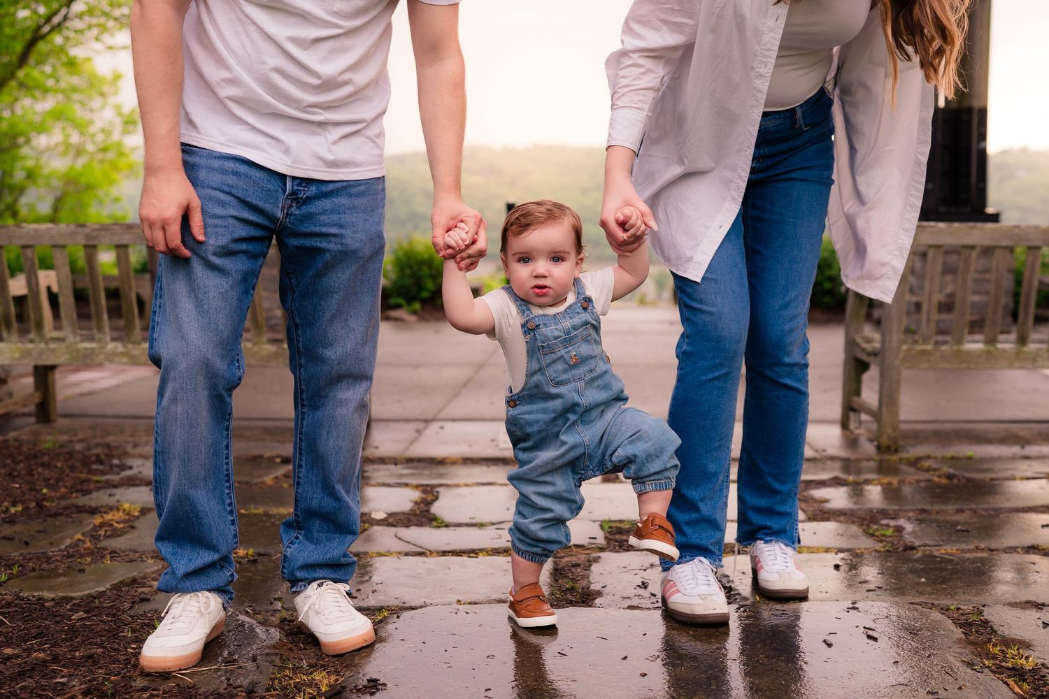 Parents helping toddler take first steps on wet pavement outdoors, baby wearing denim overalls with white sneakers.