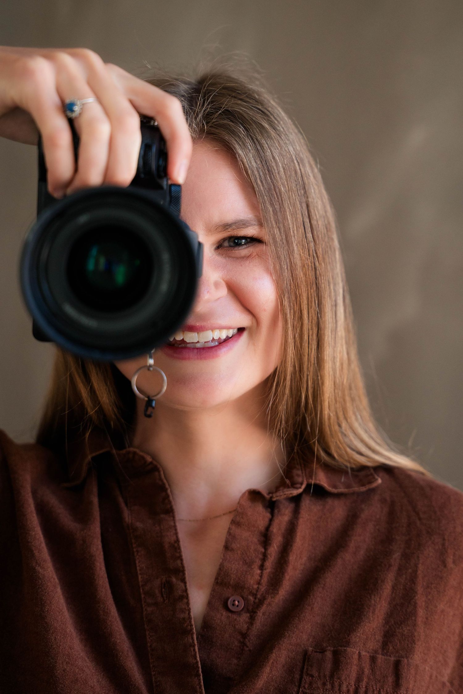 Smiling female photographer holding a DSLR camera up to her eye against a neutral background.